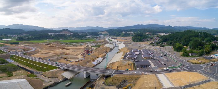 中央に川が流れる広大な工事現場に、複数の道路や橋、建物が広がり、周囲には緑の野原や丘、遠くに山々が見える様子を写した開発中の地域のパノラマ写真