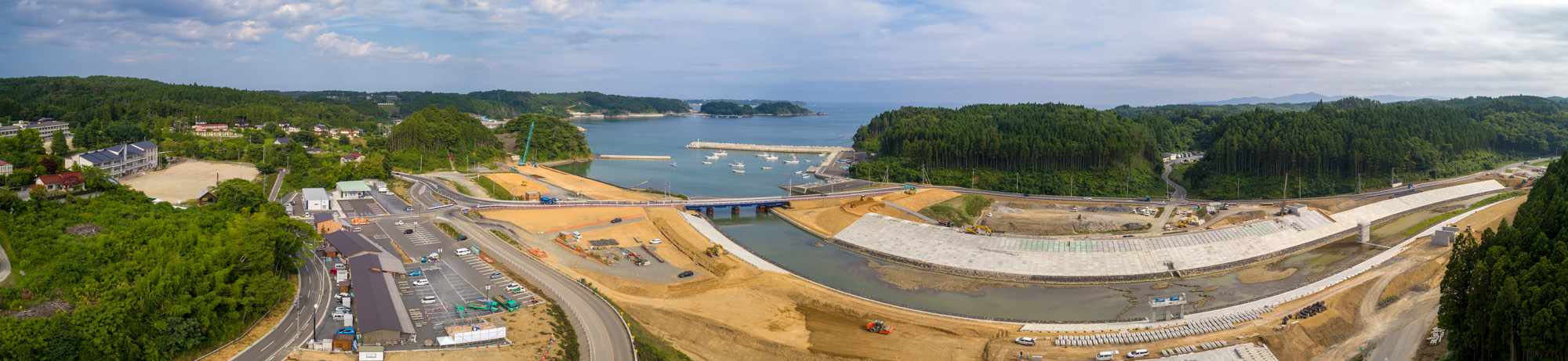 川または運河にコンクリートの護岸が整備され、橋が両岸をつなぎ、建設車両や機械が点在する工事現場の周囲に緑の丘や建物、道路、小さな港に停泊する船が見える様子を写した沿岸開発のパノラマ写真