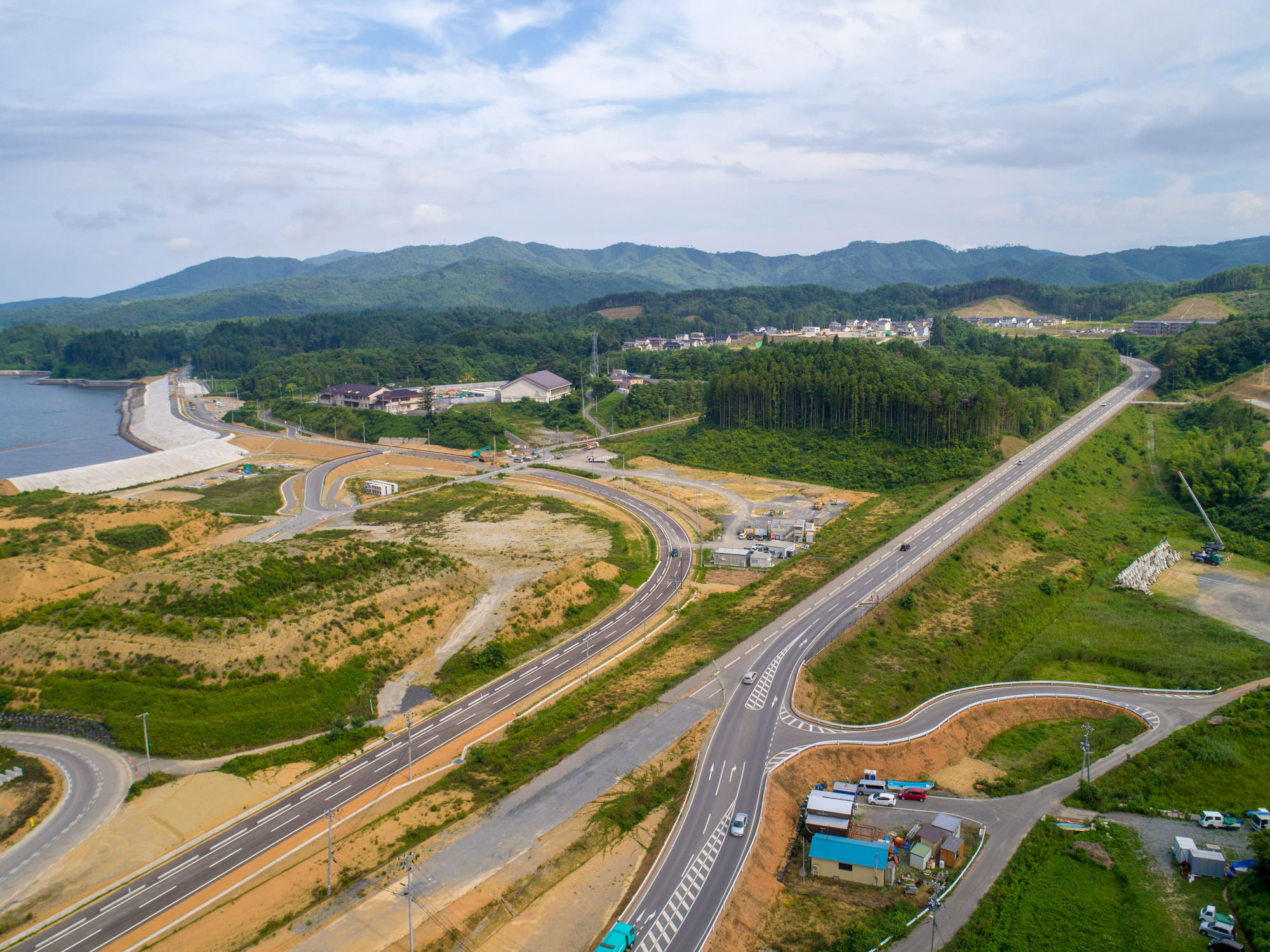 緑の野原や木々が広がる風景の中で、曲がりくねった幹線道路と複数の小道が交差し、点在する建物と遠くの山々が見える様子を写した田園地帯の写真