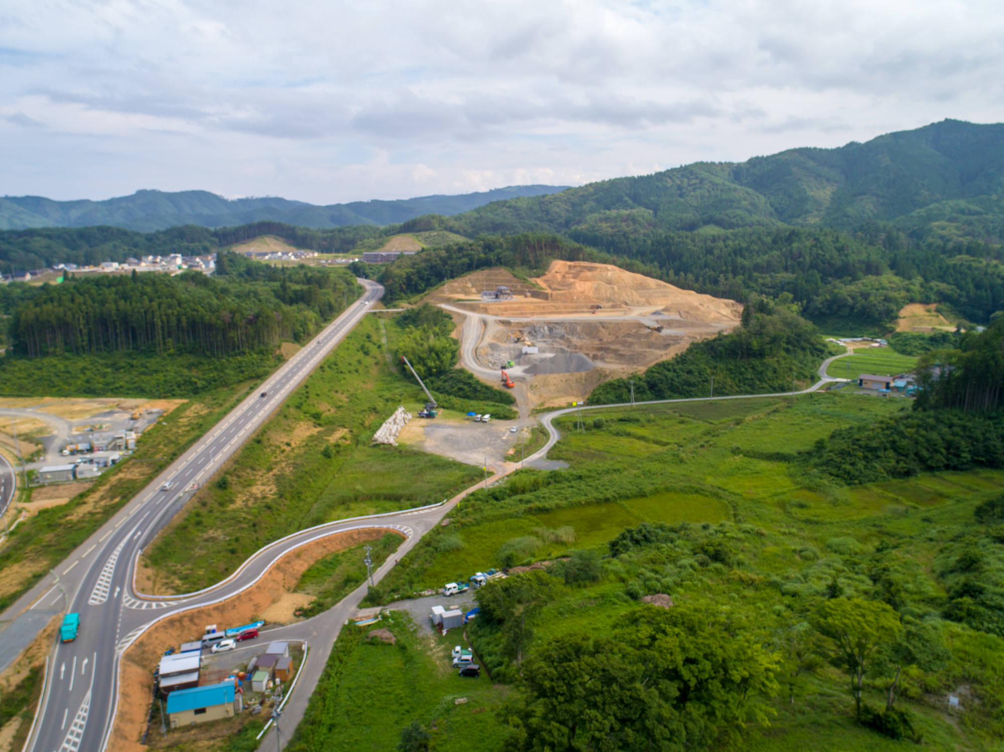 緑豊かな丘陵地に広がる工事現場で、大きく掘削されたエリアに重機や車両が見え、隣を曲がりくねった道路が通り、背景には森林に覆われた山々が広がる様子を写した山間部の開発現場の写真