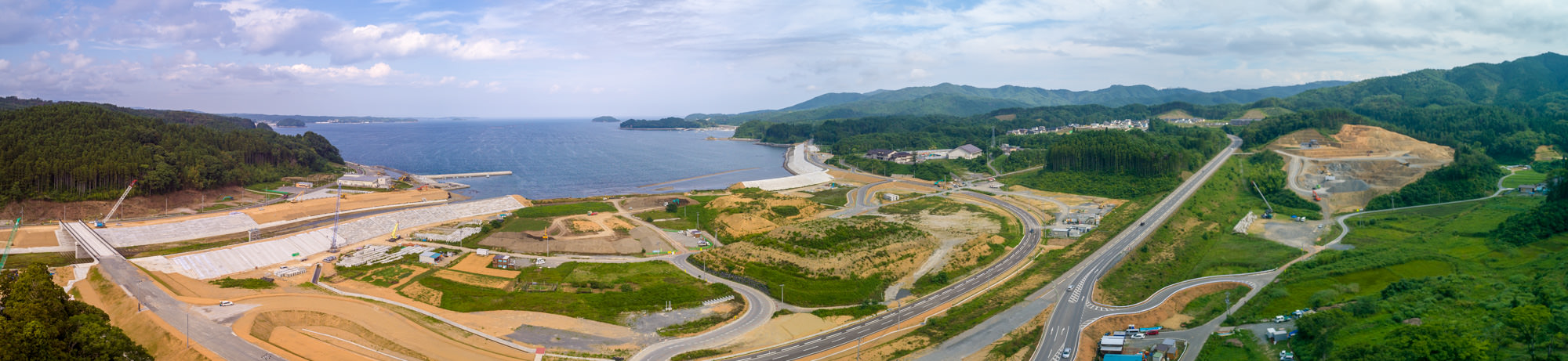 海岸沿いに広がる工事現場と、その周囲を囲む緑豊かな山々が見渡せる風景を写した海辺の開発地のパノラマ写真