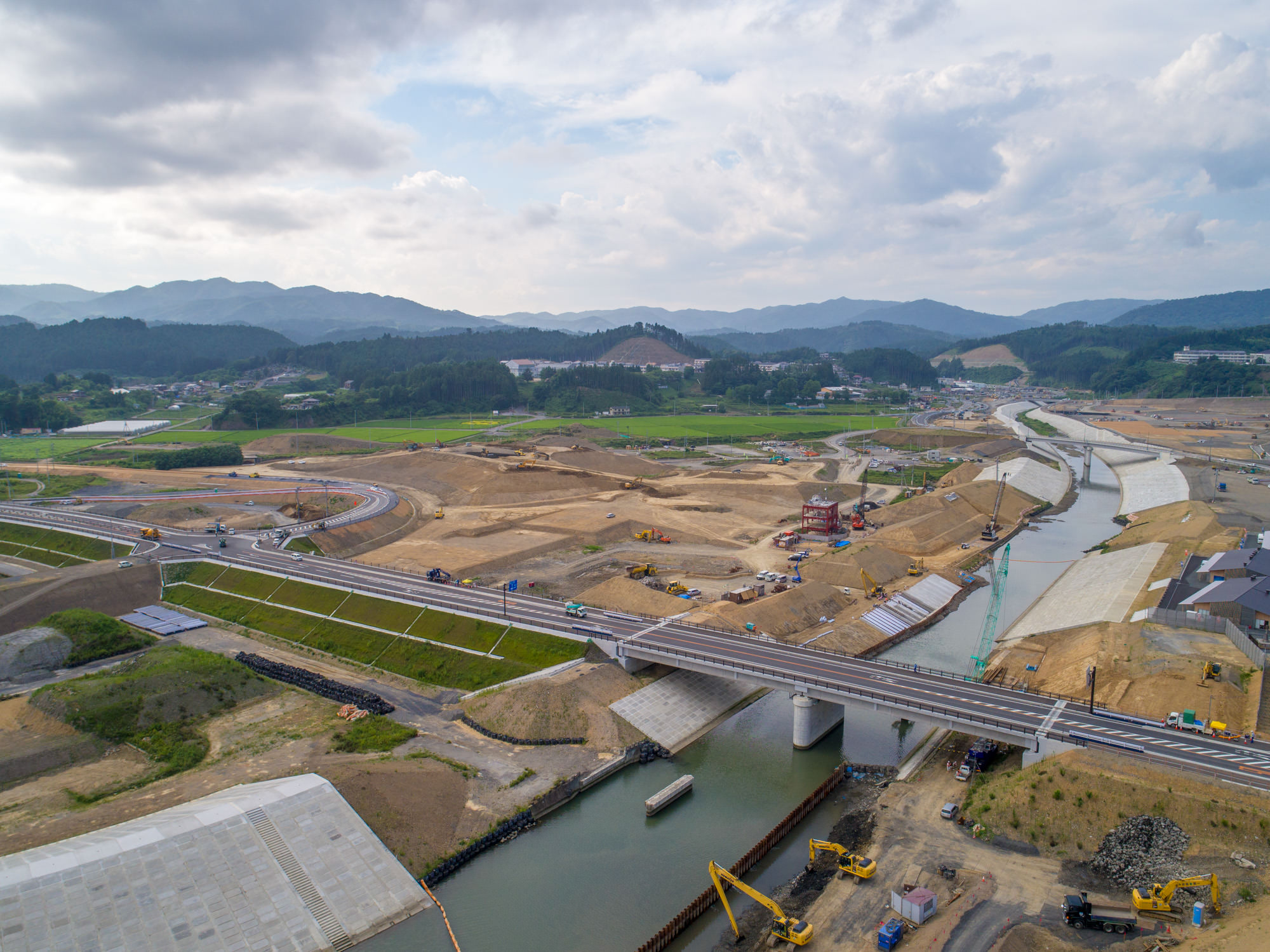 川にかかる橋を中心に、掘削機やクレーンなどの建設機械が点在し、周囲の丘陵地に道路が整備されている様子を写した山間部の橋梁工事の写真
