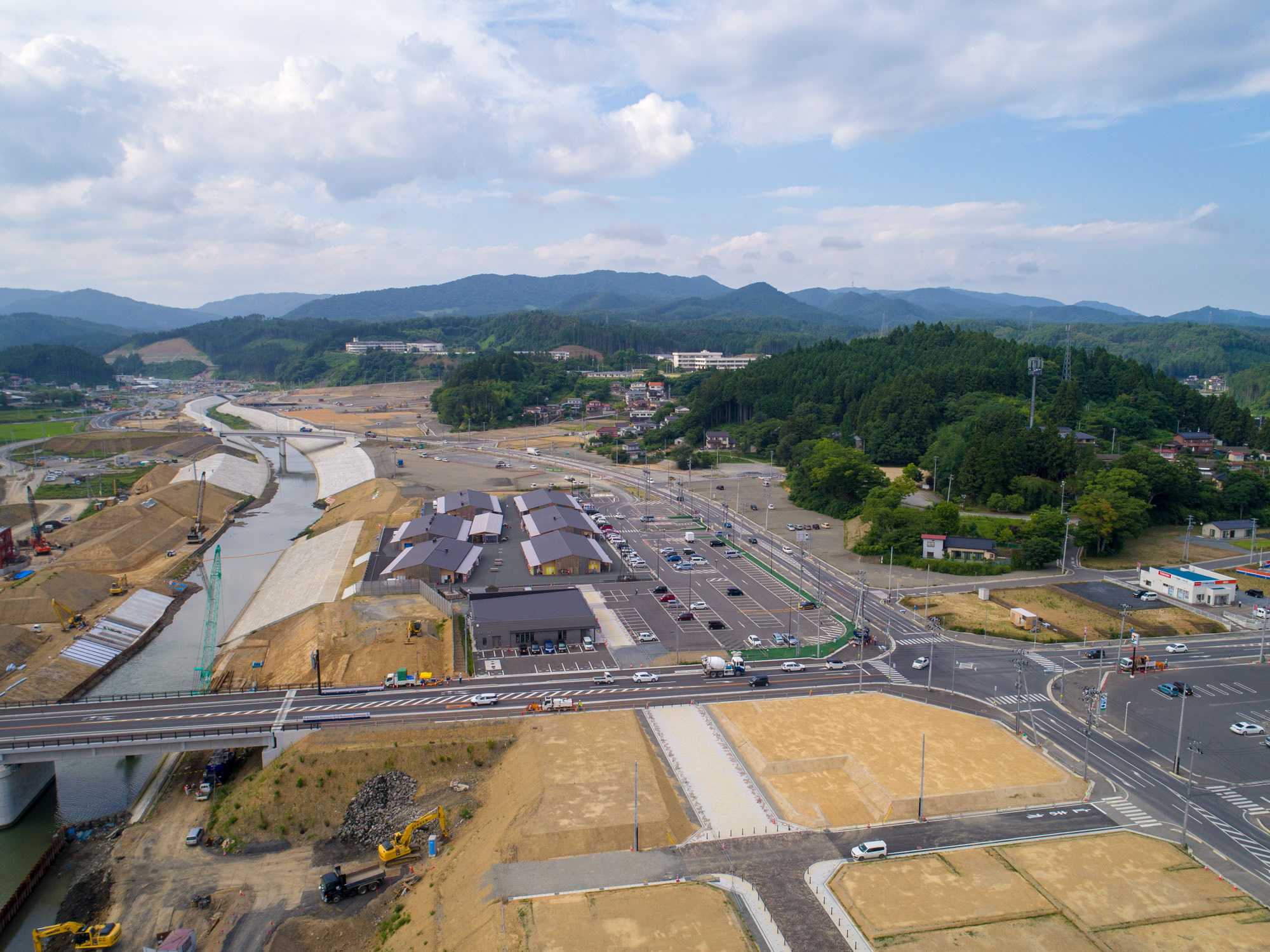 川沿いの工事現場に複数の建物や道路が広がり、周囲には丘や森林、遠くに山々が見え、建設車両や機械が点在する様子を写した山間部の開発地域の写真