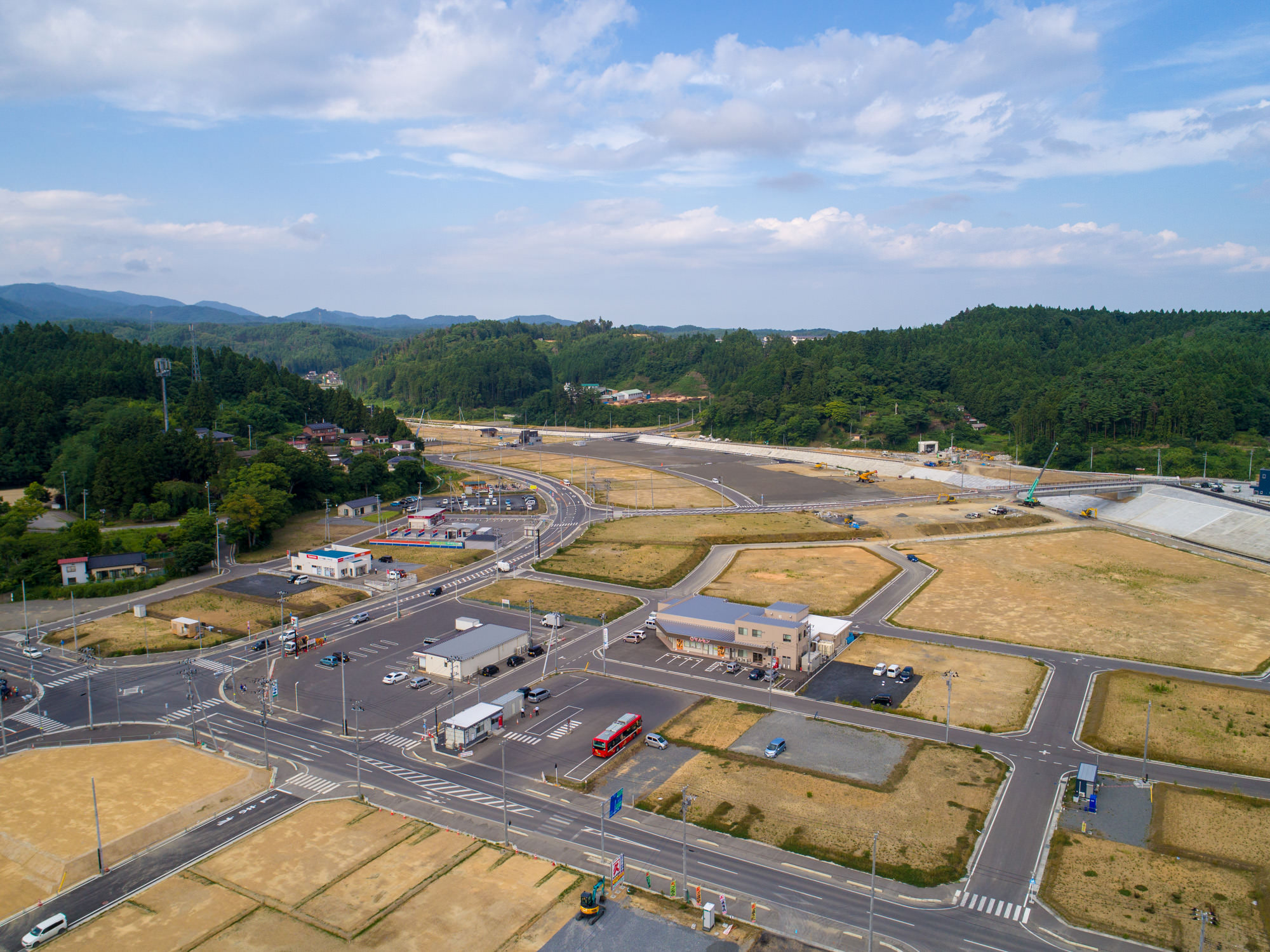 田畑や建物が点在する半農村地域を上空から捉え、交差する道路網と開発中と思われる空き地が広がり、背景には緑の丘が見える様子を写した地域開発の写真