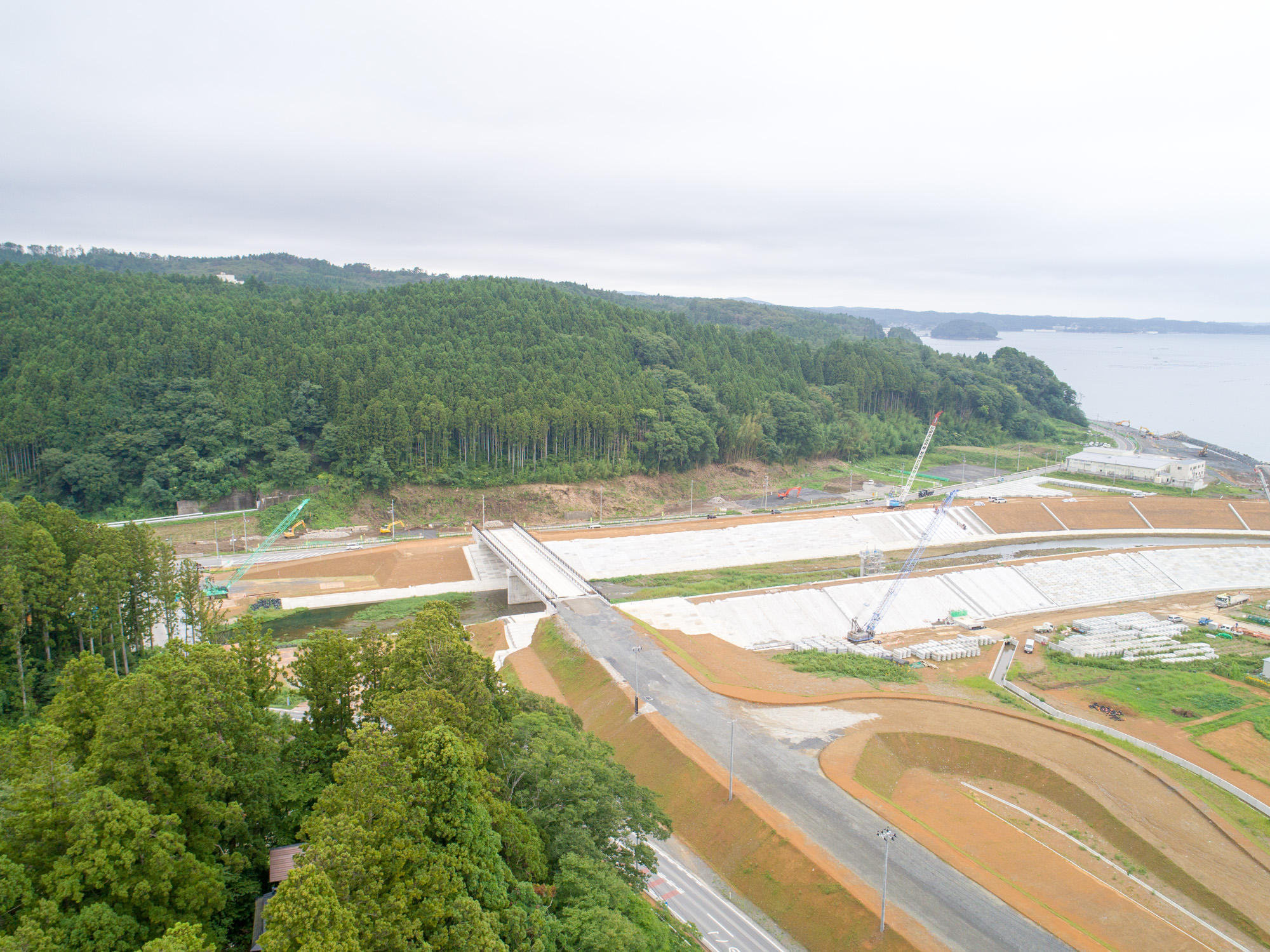 防潮堤が山の麓から海に向かって伸び、道路と橋がその上を通り、周囲には森林と重機が点在する工事区域が広がる写真