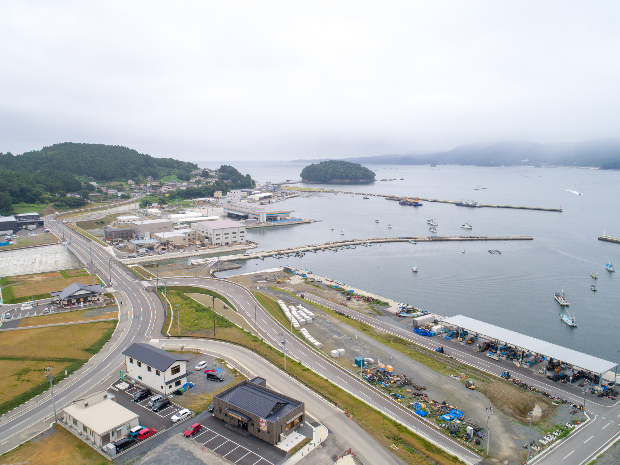 海岸沿いの町において、道路や駐車場、建物が立ち並ぶ中、橋を渡る道路工事が進められている様子が広く見渡せる写真