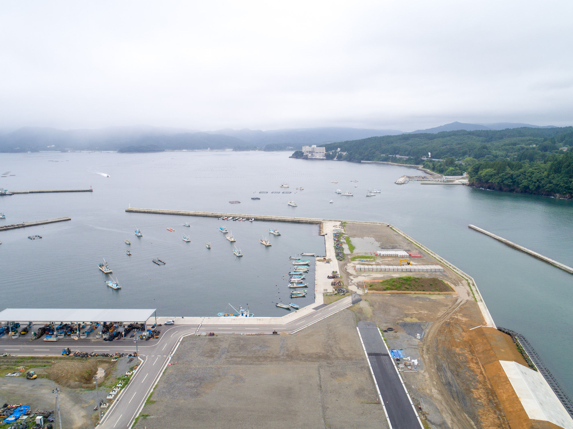 曇り空の下で複数の小型船が静かに停泊している港があり、その周囲には緑に覆われた山々と点在する建物が広がっている写真