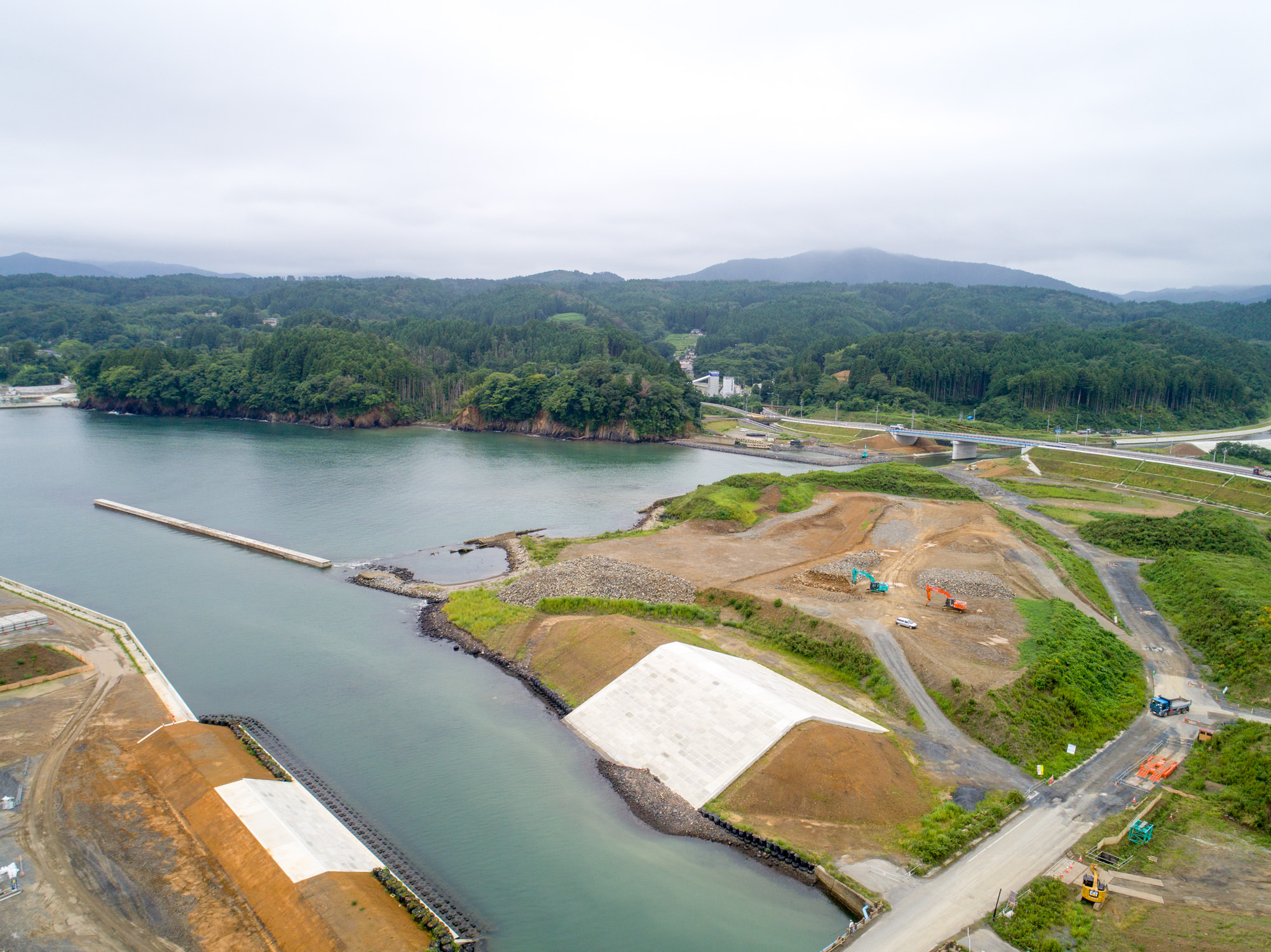 曇り空の下、湾または港と思われる水辺に面した土地が造成されており、建設資材や機材が点在する開発中の沿岸地域を上空から広く見渡し、背景には緑の木々が広がり、複数の道路や通路が敷かれている写真