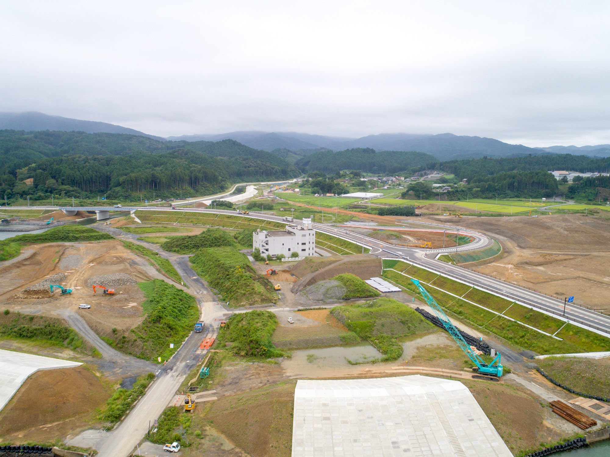 曇り空の下、中央に建設途中の建物があり、周囲には複数の道路や通路が整備されつつある工事現場が広がり、背景には森林に覆われた丘陵地と山々が見える写真