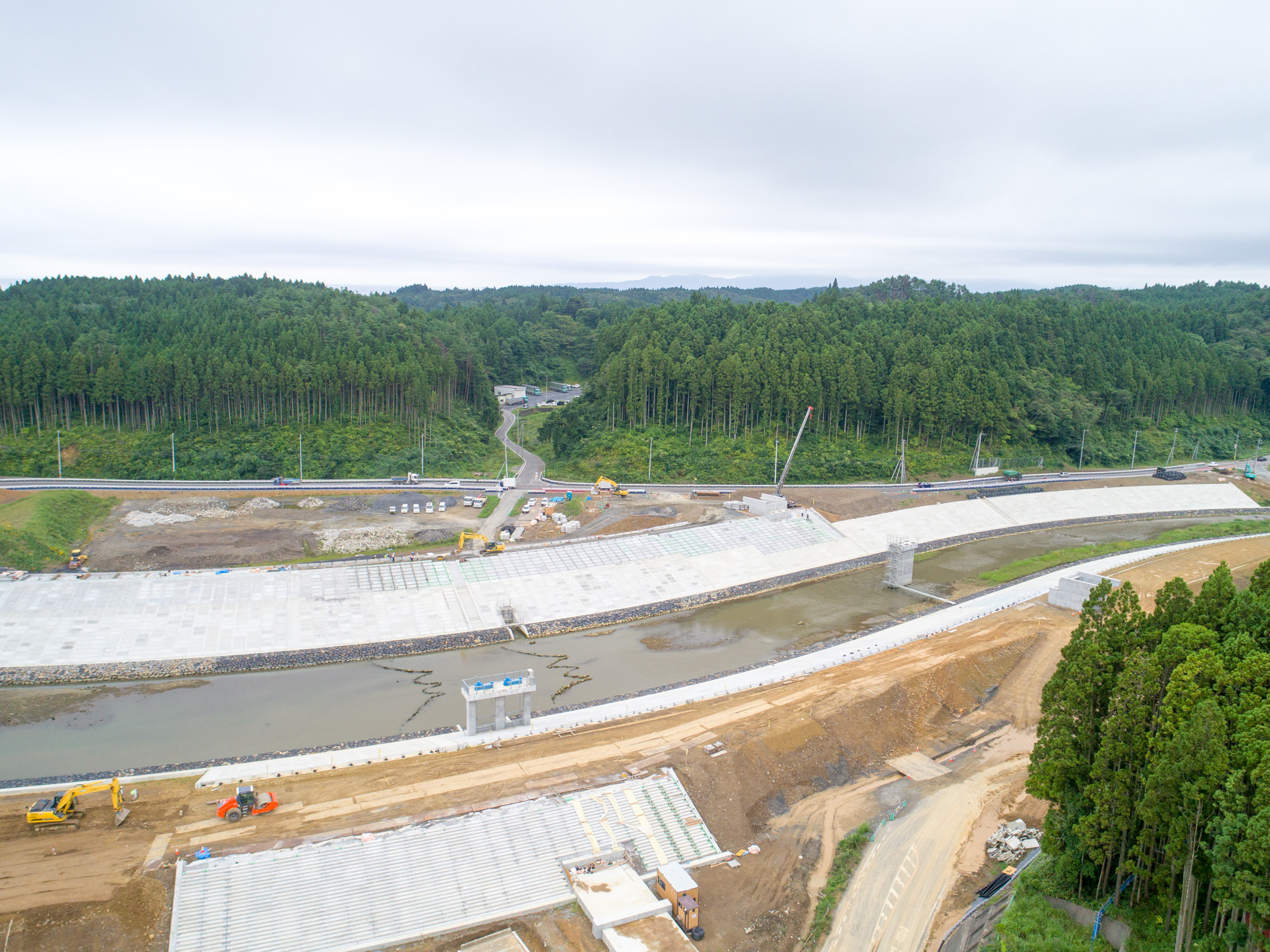 川が中央を流れる工事現場で、両岸にはコンクリート構造物が設置され、複数の建設車両や機材が稼働しており、周囲は深い森林に囲まれ、背景には森へと続く道路が見える写真