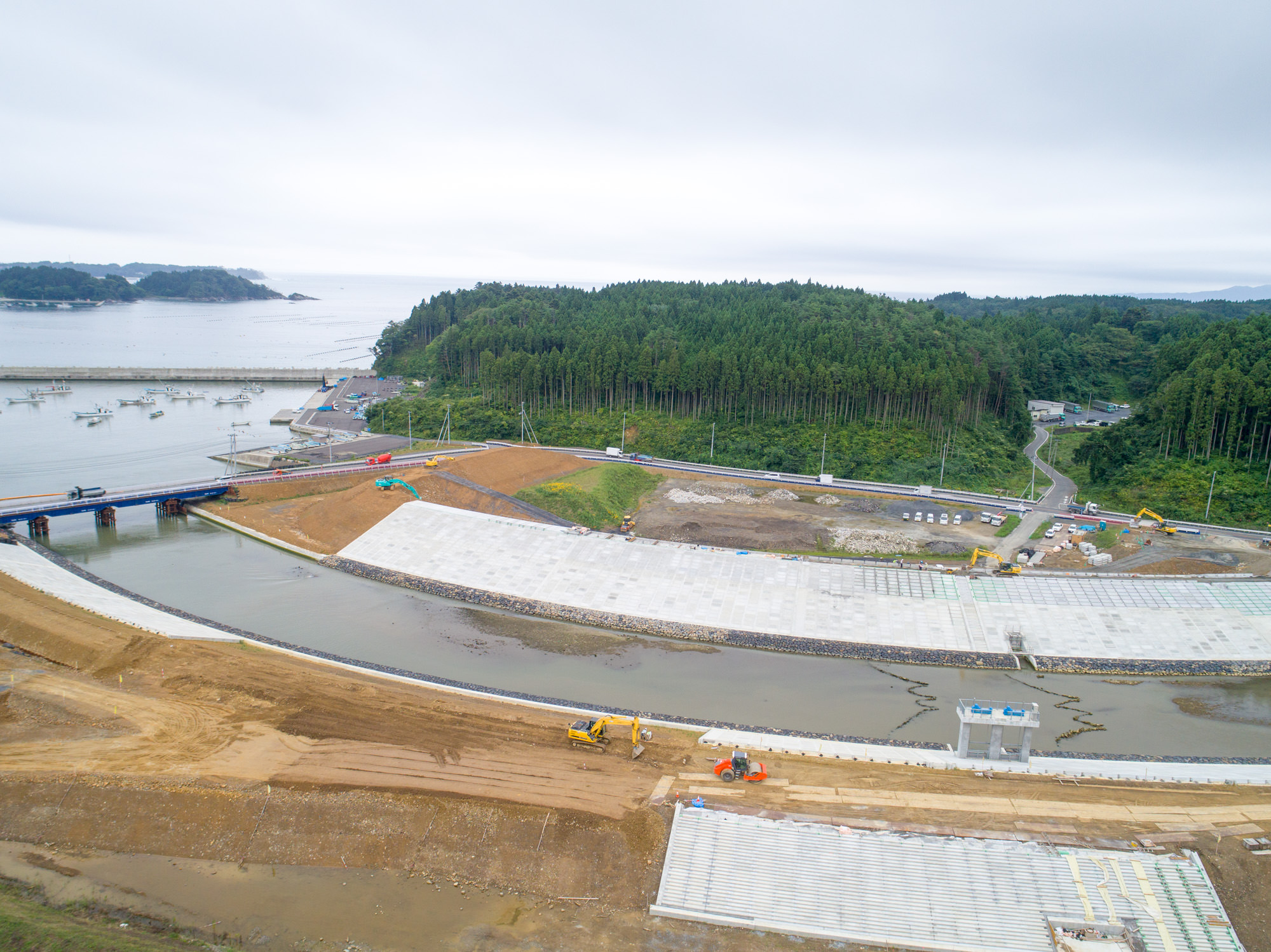 川や運河が流れる海沿いの工事現場で、複数のショベルカーなどの建設機械が稼働しており、周囲には深い森林が広がり、背景には海と思われる水面が見える写真