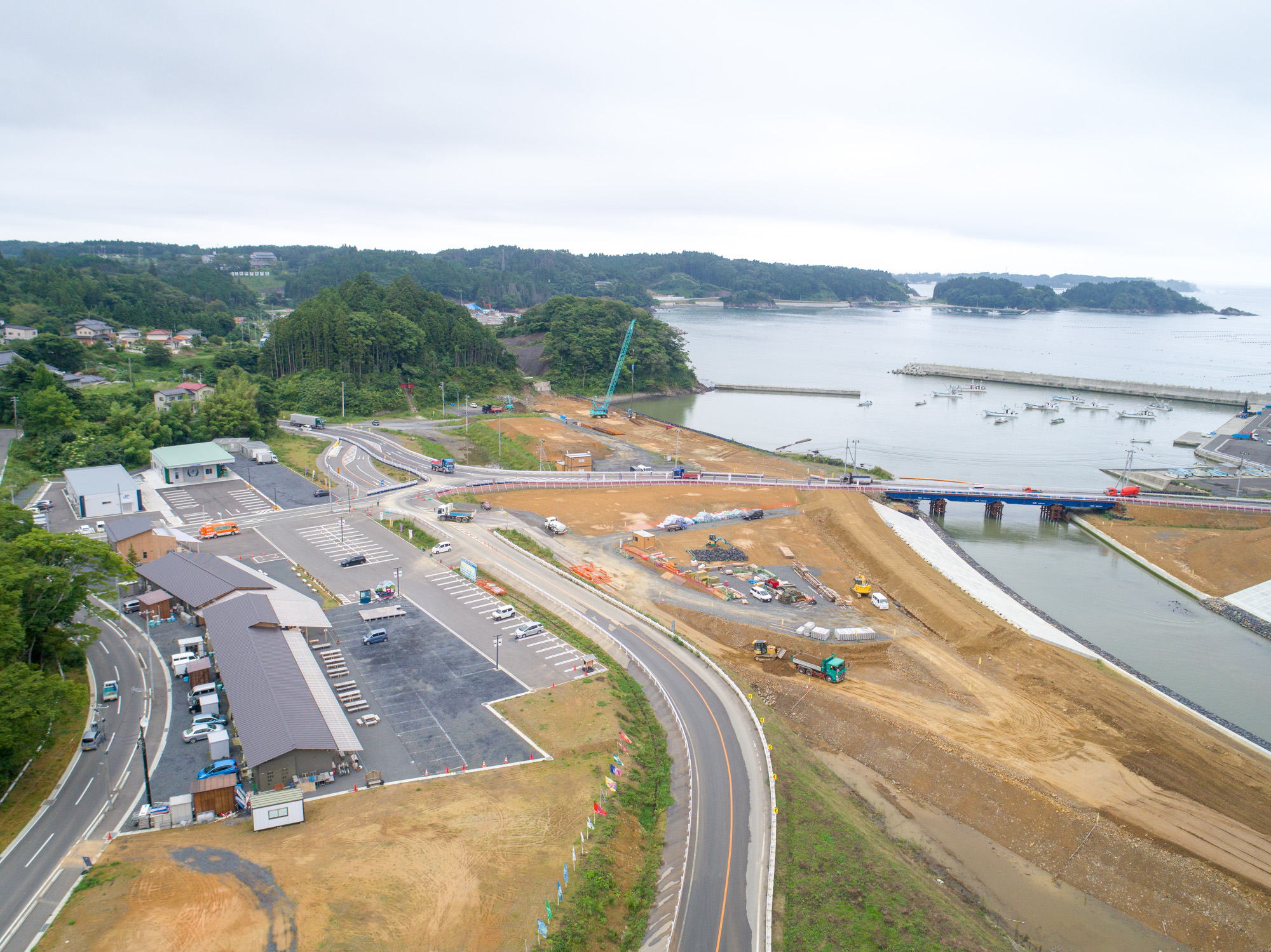 海岸沿いの町において、道路や駐車場、建物が立ち並ぶ中、橋を渡る道路工事が進められている様子が広く見渡せる写真