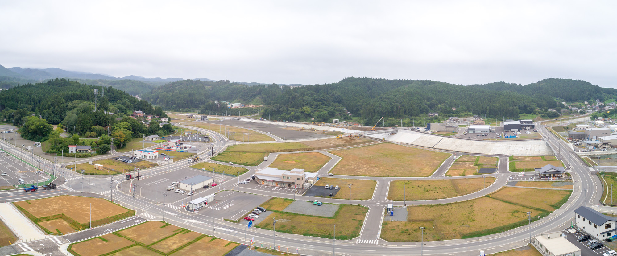 道路や区画が整備され始めており、広い空き地の中にポツポツと建物が建ち始めている様子と、周囲を囲む山や森が見える、再開発中の地域を広角で写した写真