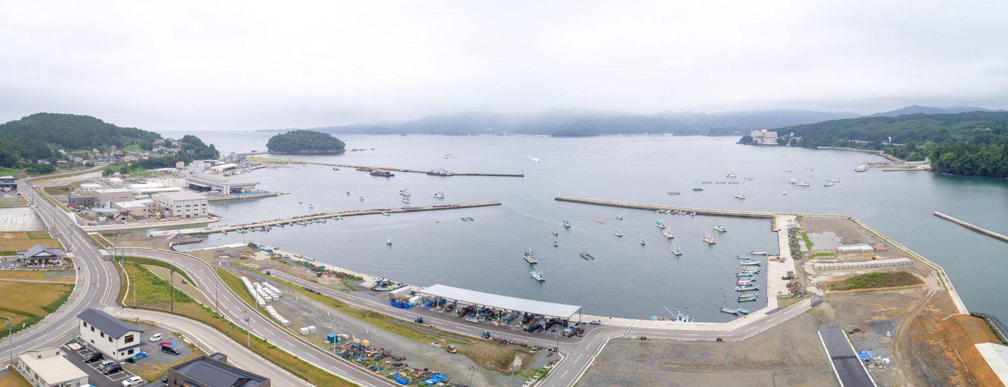 川沿いの工事現場では重機が稼働しており、その周囲には緑豊かな森林が広がり、さらに奥には青く穏やかな海が見える風景が広がっている写真