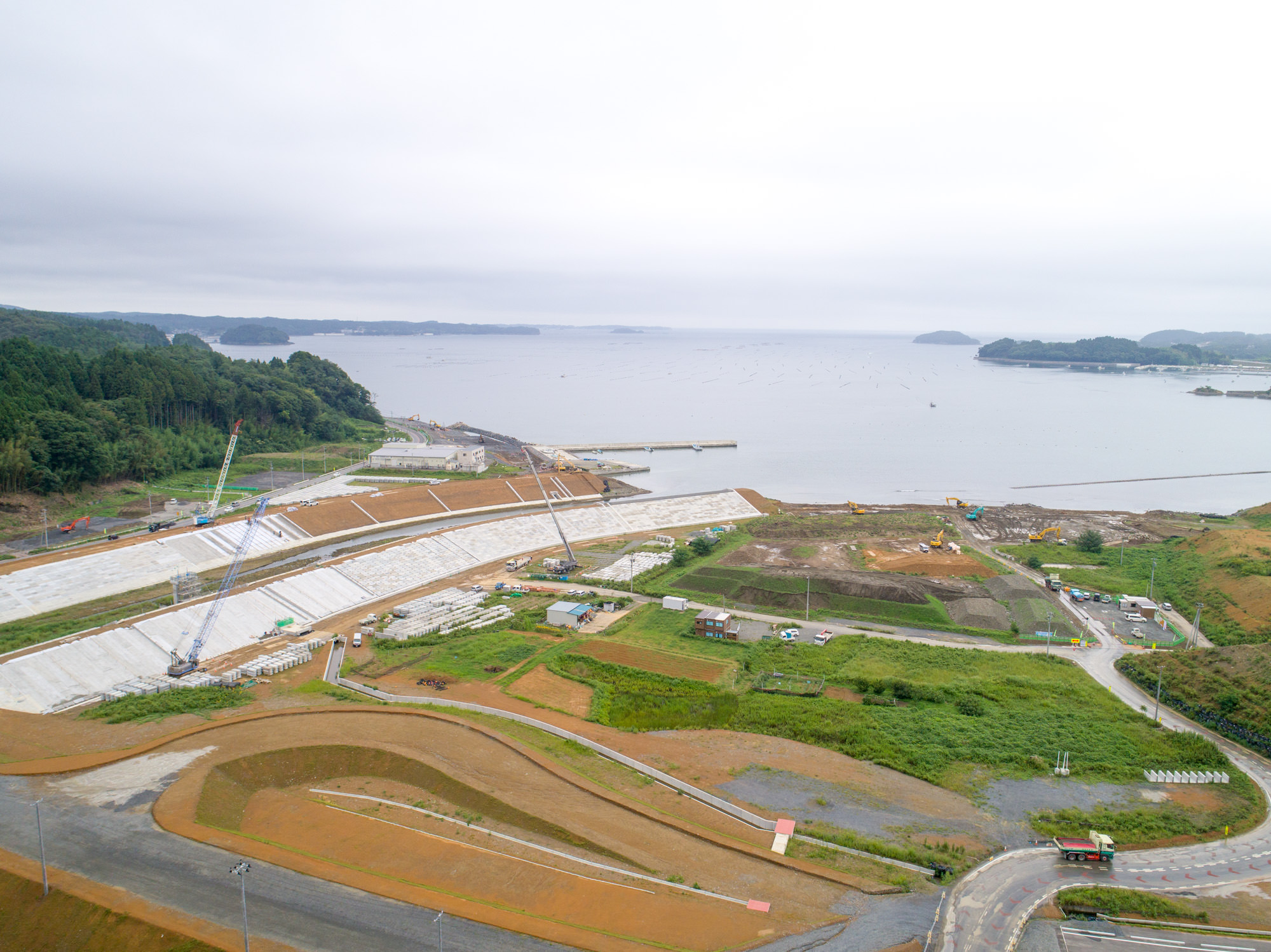 防潮堤が海岸線に沿って築かれ、その内側には重機が作業を進める工事区域や道路、植生の残る土地が広がる写真