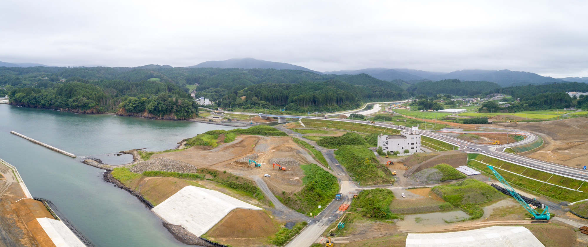 曇り空の下、川または湖の近くに広がる工事現場ではクレーンや車両などの建設機材が点在し、複数の道路や通路が敷かれ、背景には緑の丘が広がっている写真