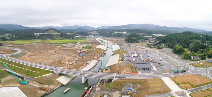 川沿いの谷間にかかる橋と、周囲で進む道路や建物の工事現場、そして山々に囲まれた地域全体を見渡す構図の写真