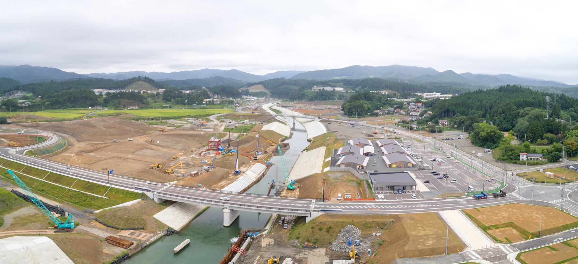 川に沿って堤防工事が進められており、大型重機やクレーンが作業している様子と、新しく整備された道路や橋が山あいの町を横切っている様子が見える、高台からの広範囲を写した復興中の街並みの写真