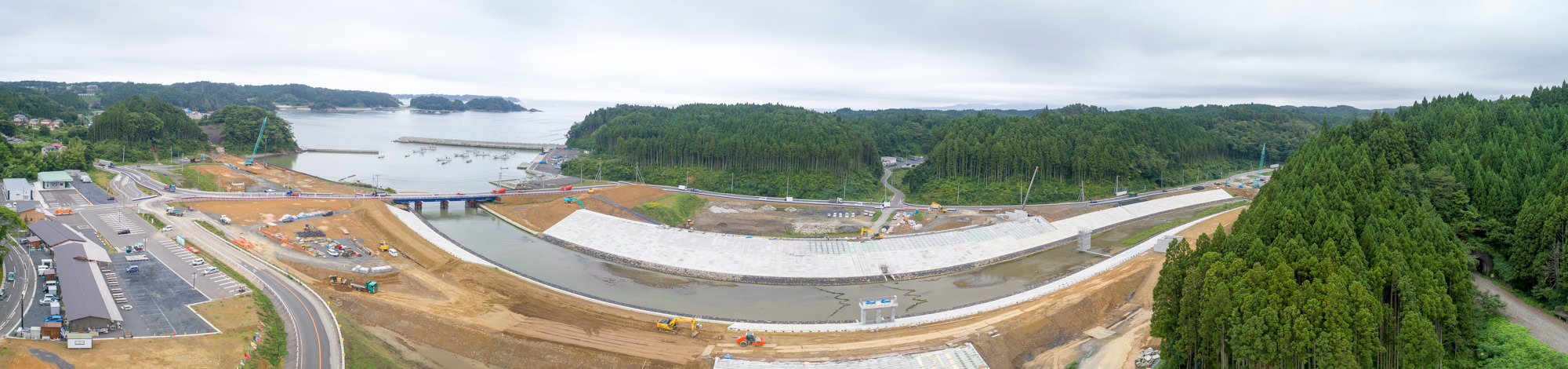 川沿いの工事現場では重機が稼働しており、その周囲には緑豊かな森林が広がり、さらに奥には青く穏やかな海が見える風景が広がっている写真