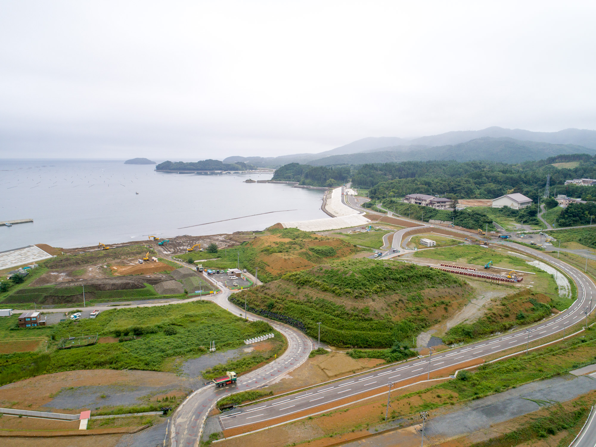 湾に沿って築かれた防潮堤と、そこへ続く新しい道路網、その周囲に広がる整地された工事現場や建物が見える写真