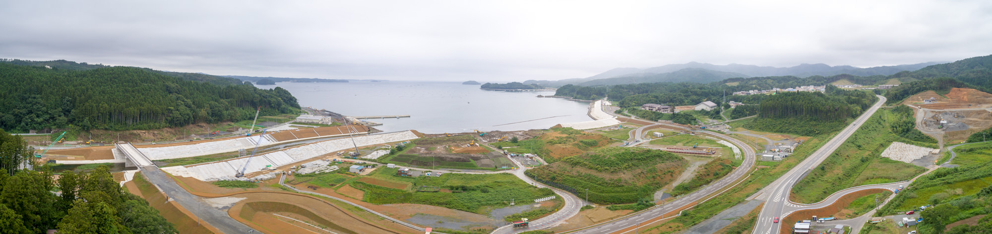 パノラマ海に面した湾岸エリアで、大規模な防潮堤や道路の建設が進められており、背後には山並みが広がる風景の写真