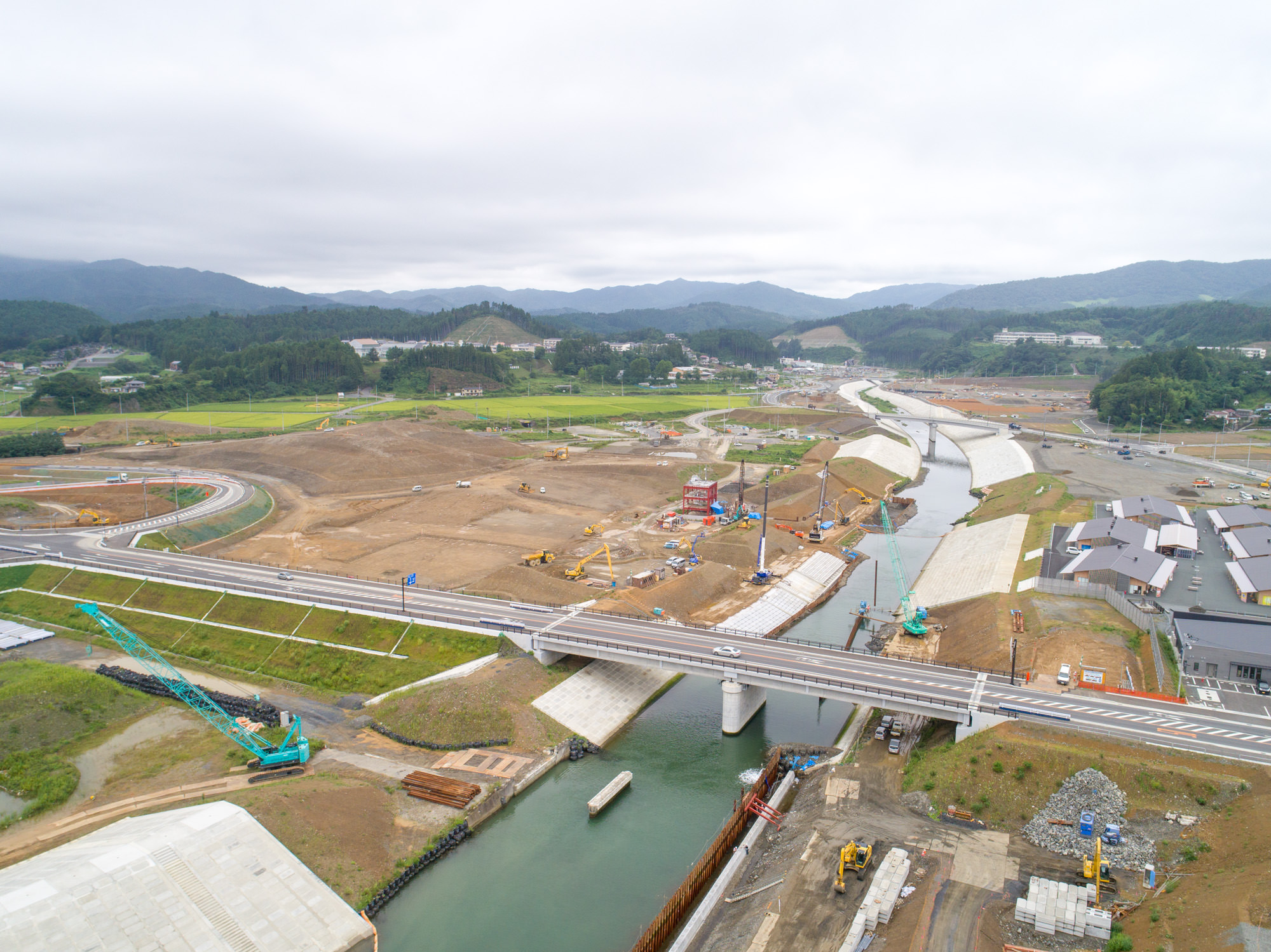 川に架かる橋の下で工事が行われており、重機やクレーンが川沿いに並び、背景には田畑と山々が広がる、災害復興の様子を俯瞰で捉えた写真