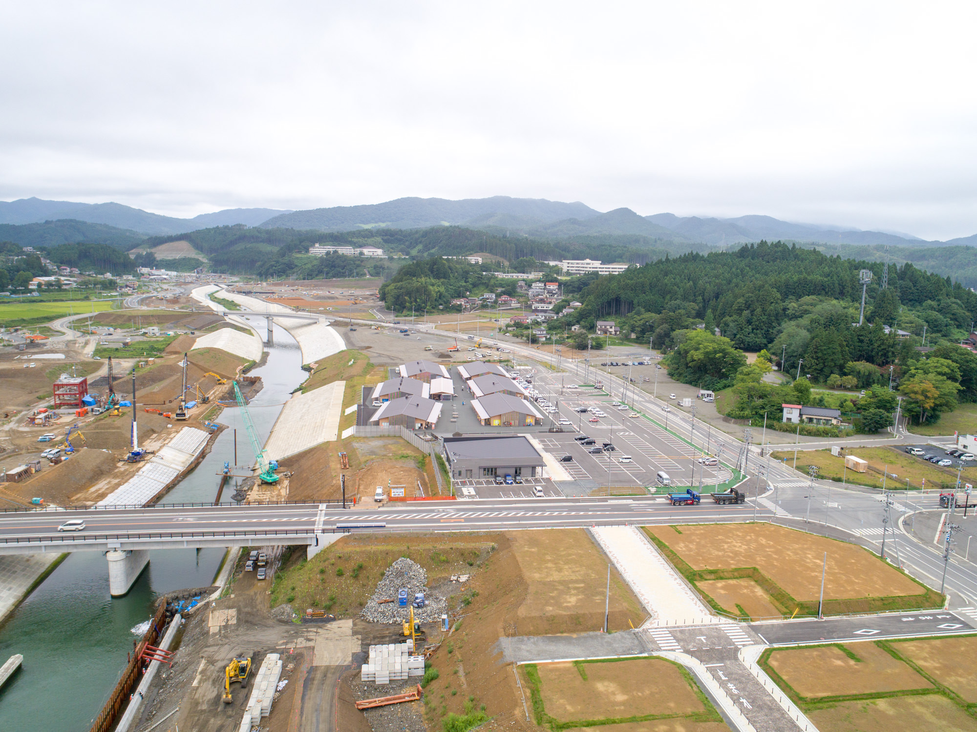 川の右側に整備された建物群と広い駐車場が並び、川沿いの護岸工事やその奥のまちづくりが進んでいる様子を高所から見渡した写真
