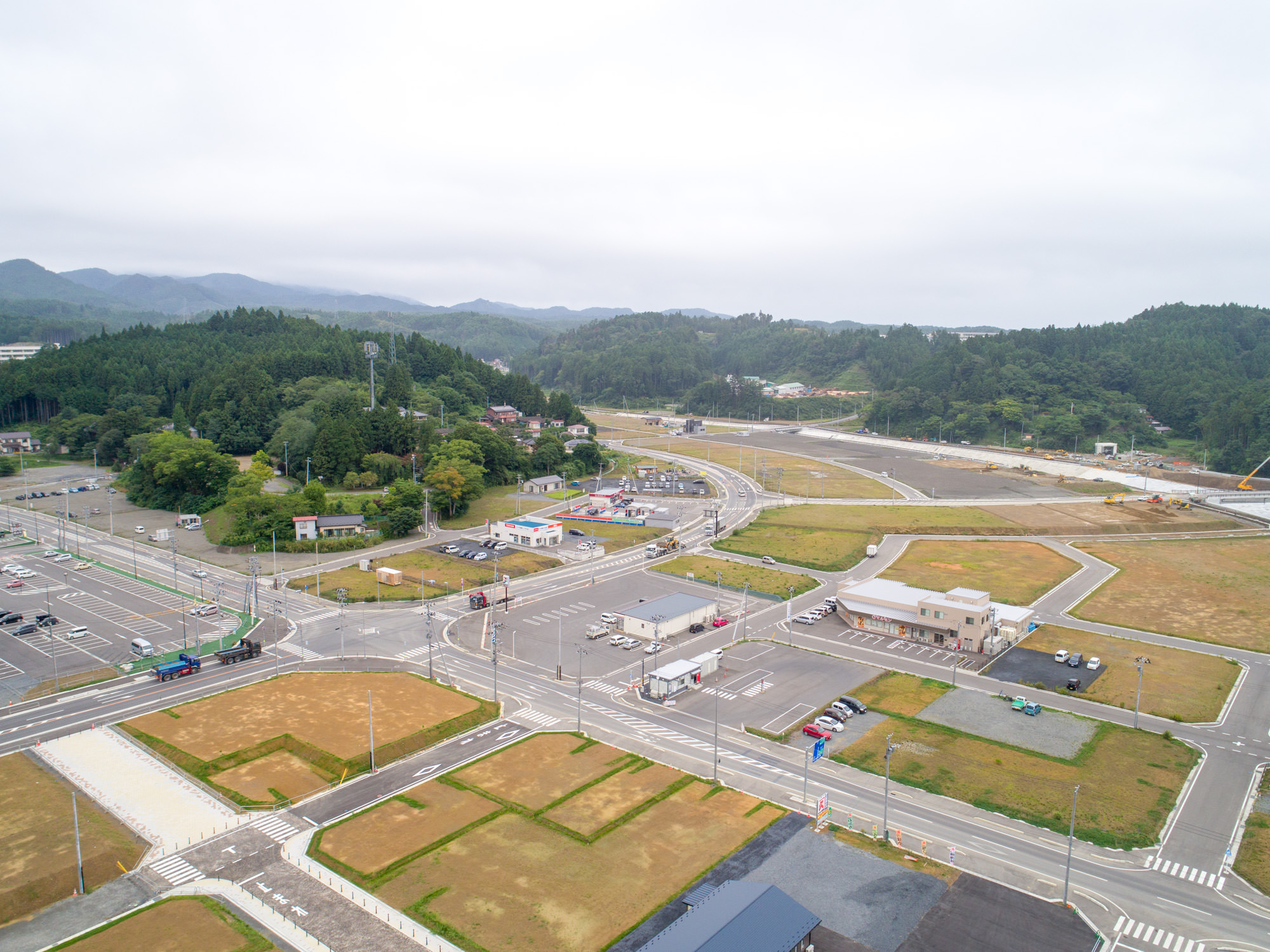 森のふもとにある小さな建物やガソリンスタンドのある交差点を中心に、周囲の整備された道路や区画、復興途中の広い敷地が写っている、再生中の街区の写真