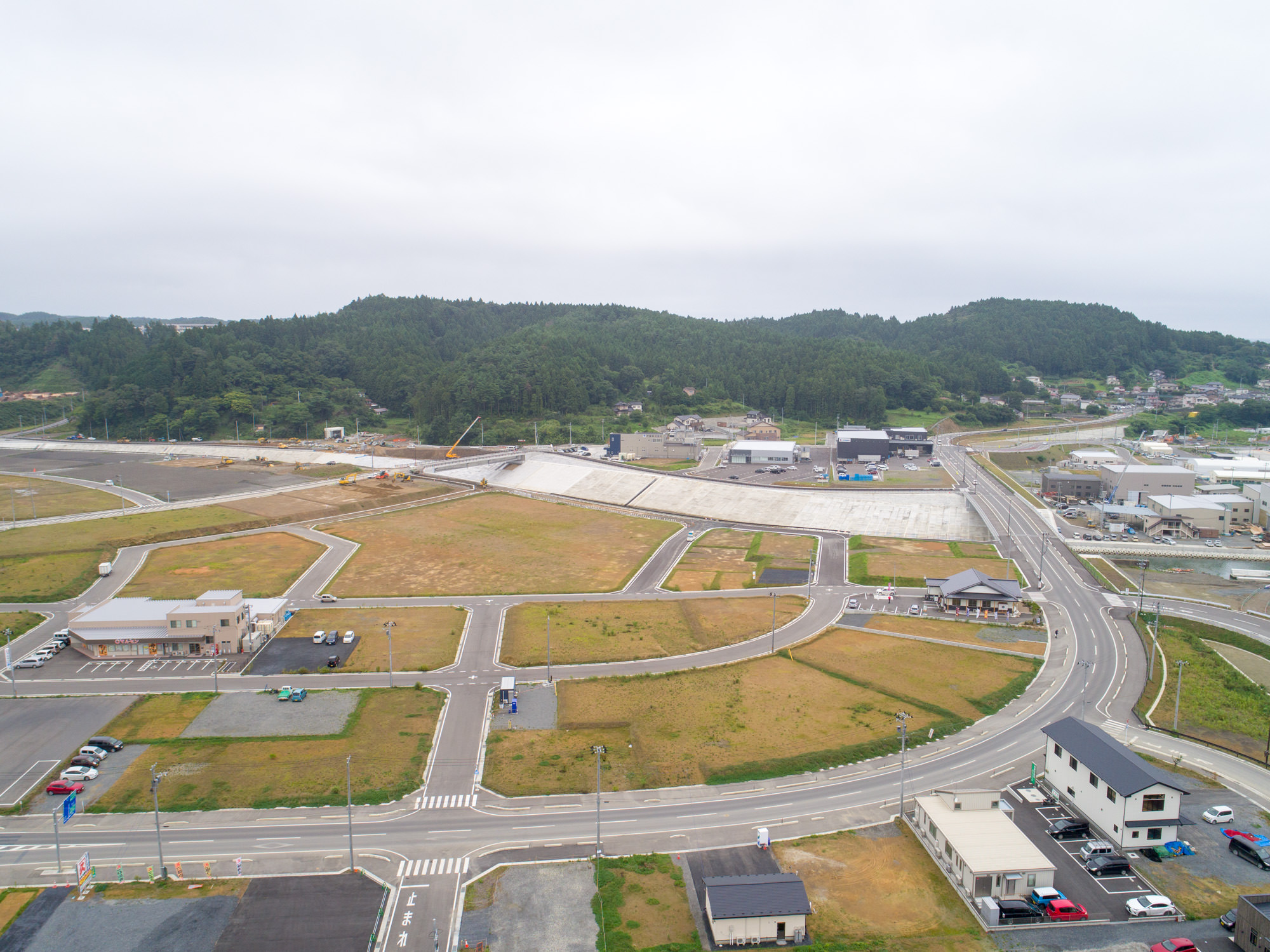 右手に川と防潮堤、左手に広く整地された区画と建物が数棟建ち始めている様子を、交差点とカーブした道路越しに見渡した復興エリア全体の写真