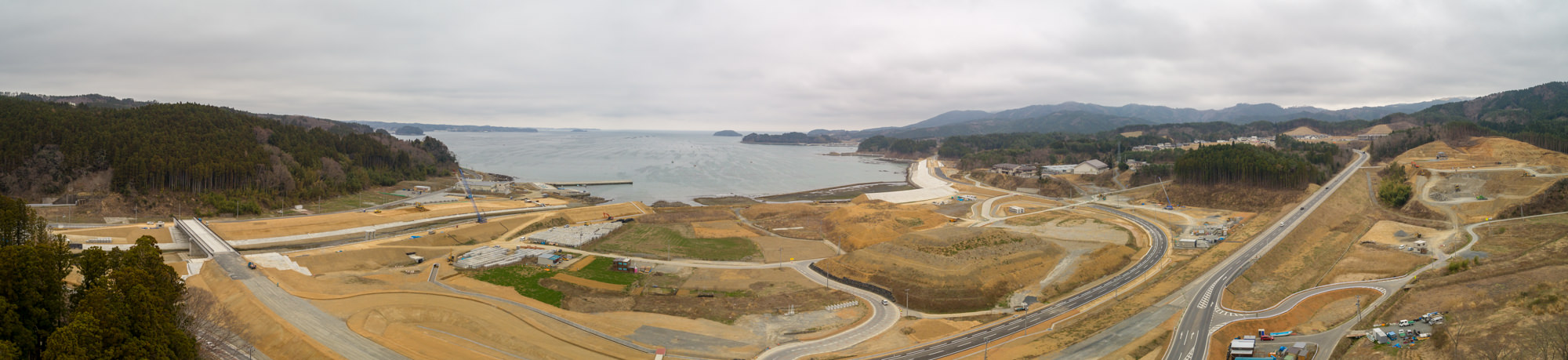 曲がりくねった道路や複数の建設現場、緑地が点在する海岸地域をパノラマで捉えた写真で、背景には海が地平線まで広がり、周囲には丘や森林が広がり、曇り空が空一面を覆っている静かな風景の写真