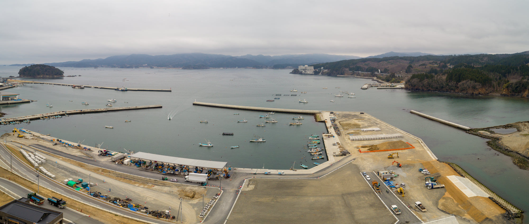 曇り空の下、港に複数の船が停泊し、防波堤が海に向かって伸びる沿岸地域をパノラマで撮影したもので、周囲にはクレーンやトラックなどの建設機材が見られ、背景には丘や山々が広がる風景の写真