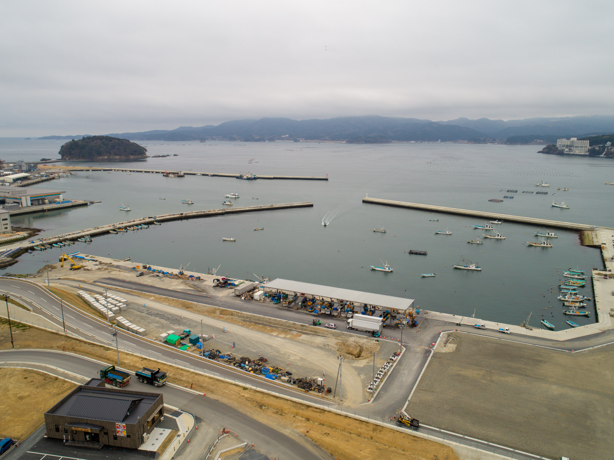 曇り空の下、港に複数の船や船舶が停泊し、防波堤で囲まれた沿岸地域を上空から撮影したもので、岸沿いには倉庫のような建物や構造物が並び、周囲には道路や工事中の区域、小さな島が遠くに見える風景の写真