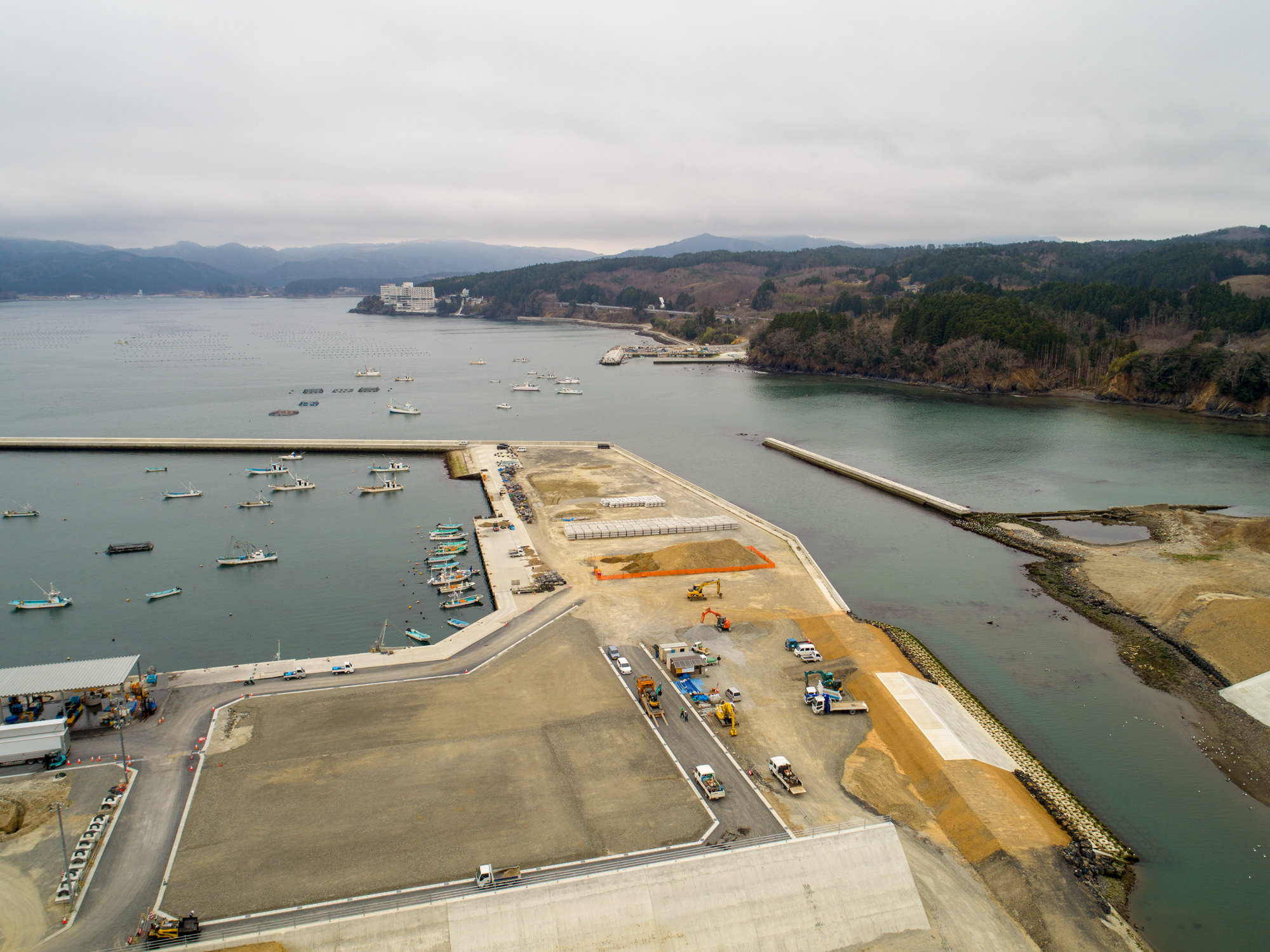 曇り空の下、港に複数の船が停泊し、防波堤に囲まれた沿岸地域を上空から撮影したもので、陸地では建設作業が行われており、建物や車両が見られ、背景には丘や森林が広がる風景の写真