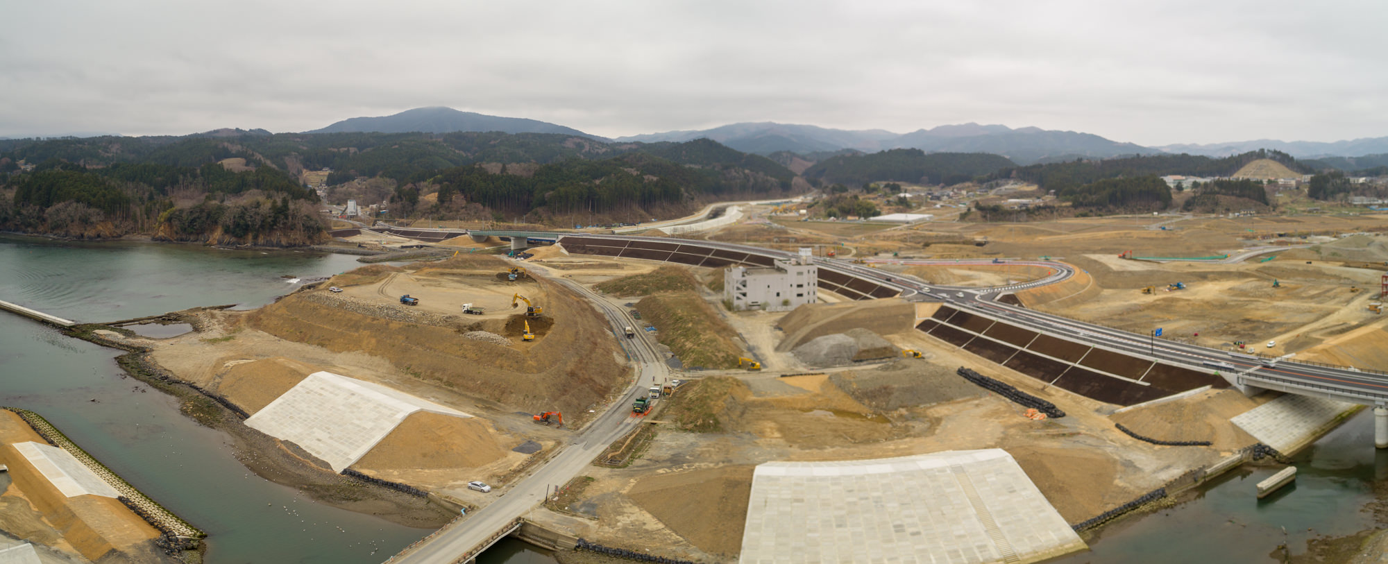 曇り空の下、川沿いに広がる大規模な建設現場を上空から撮影したもので、複数の道路や橋が建設中であり、重機や車両が稼働していて、背景には丘や山々、一部に植生が見られる風景の写真