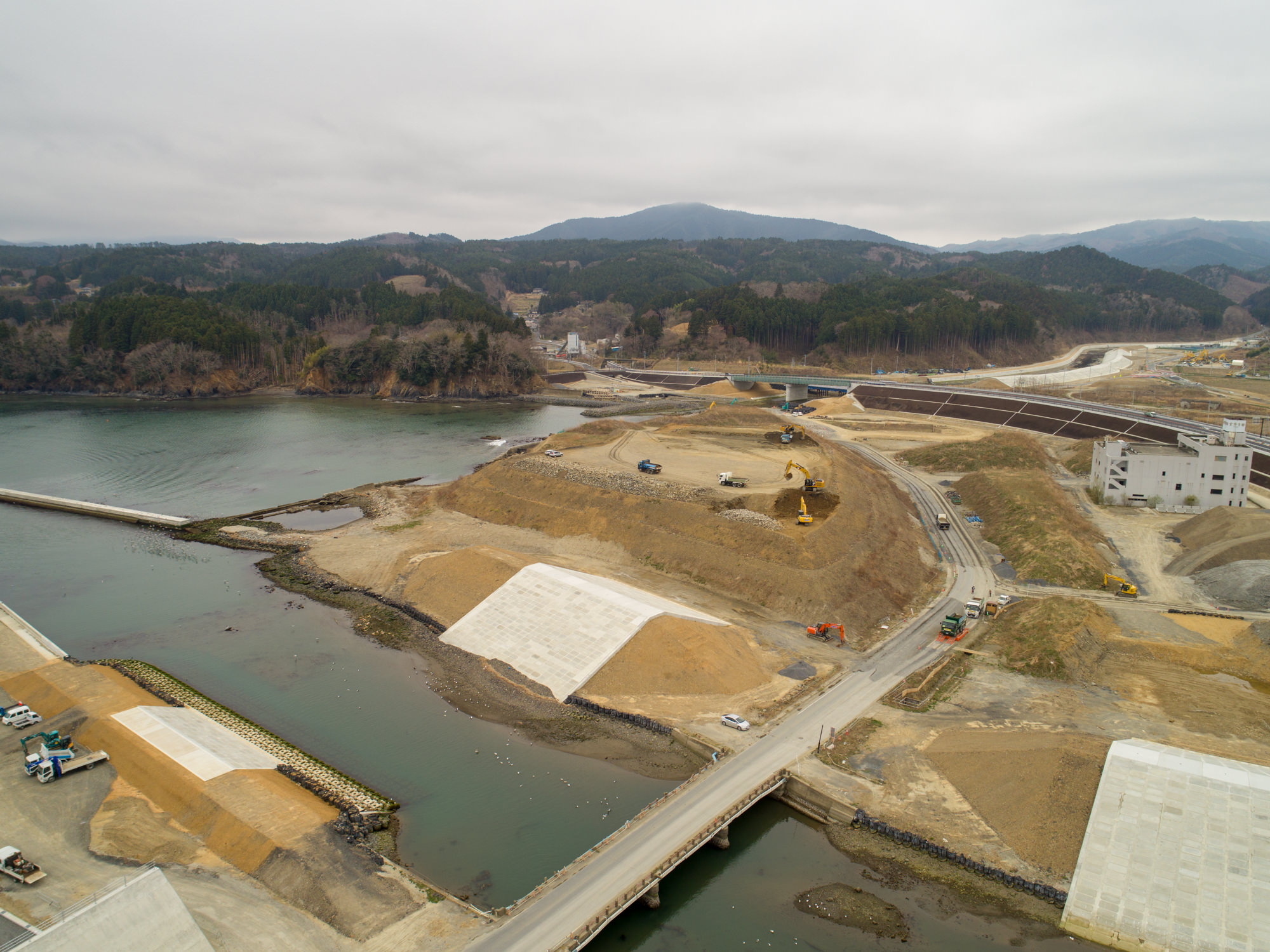 曇り空の下、川沿いにある建設現場を上空から撮影したもので、複数の土盛りとコンクリート補強が施された部分があり、水上には橋が架かって現場をつないでおり、土盛りや道路上には建設車両や機材が見られ、背景には森林に覆われた丘や山々が広がる風景の写真