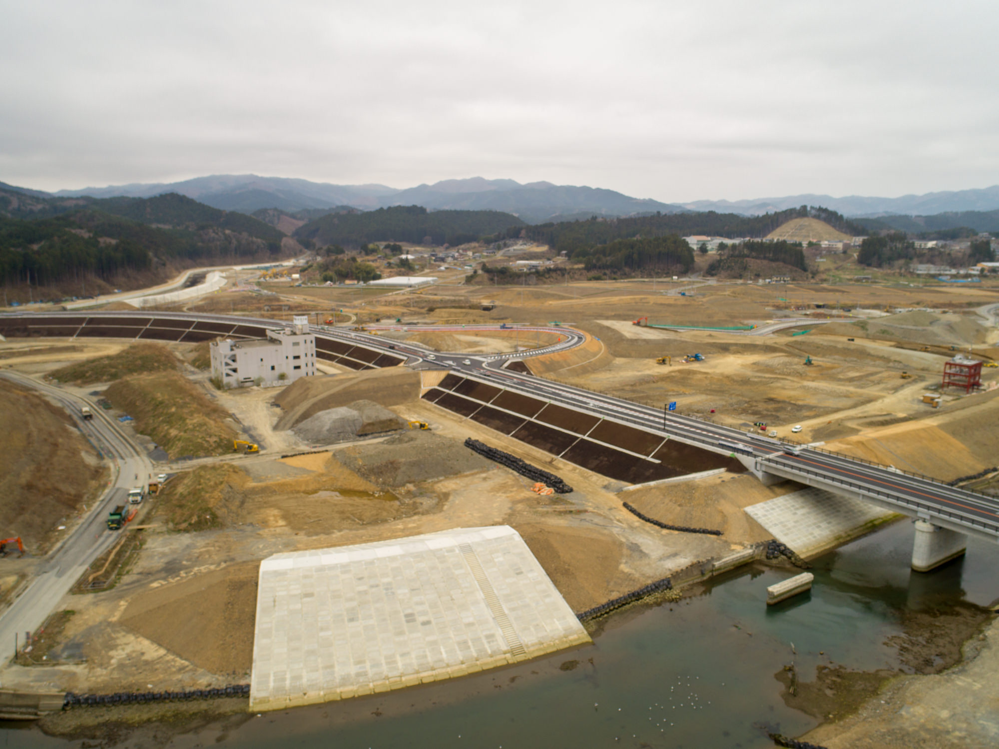 曇り空の下、水辺に架かる橋を中心とした建設現場を上空から撮影したもので、道路や土工事、一部の構造物が見られ、背景には丘や山々が広がる風景の写真