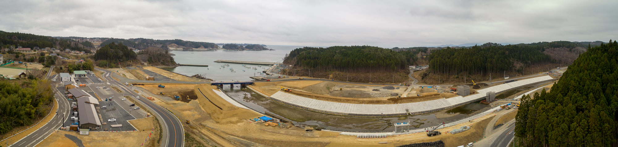 曇り空の下、左側に道路や駐車場、建物が並び、中央にはコンクリート護岸と橋が架かる川が流れ、背景には船や桟橋がある湾と森林に覆われた丘が広がる、開発または工事中の沿岸地域をパノラマで撮影した風景の写真