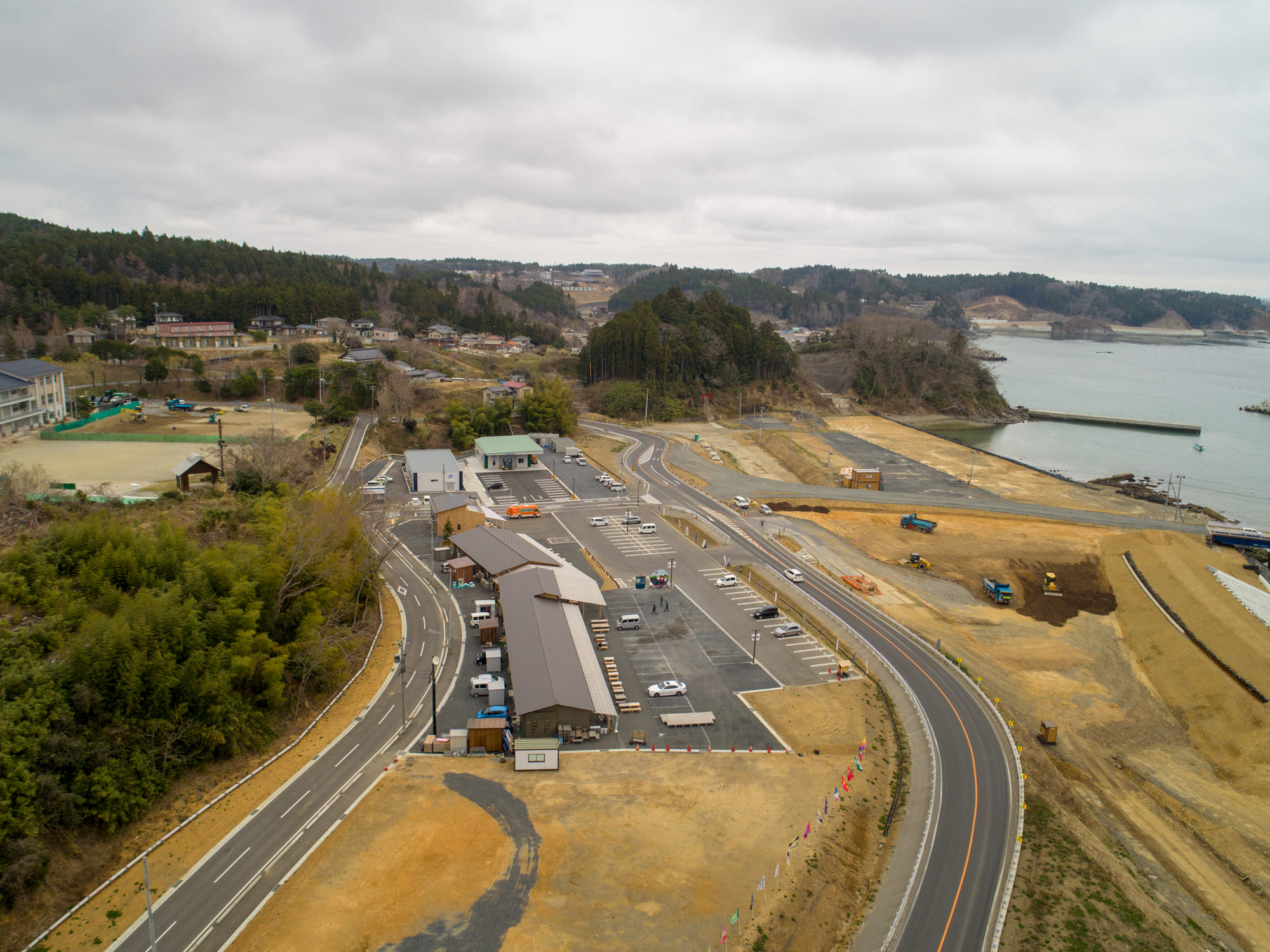 曇り空の下、右側に海岸線が広がる沿岸地域を上空から撮影したもので、湾曲した道路が複数の車が停まっている駐車場へと続き、右側の海岸付近では建設作業が行われている建設車両や機材が見られ、背景には木々に覆われた丘と点在する住宅が広がる風景の写真