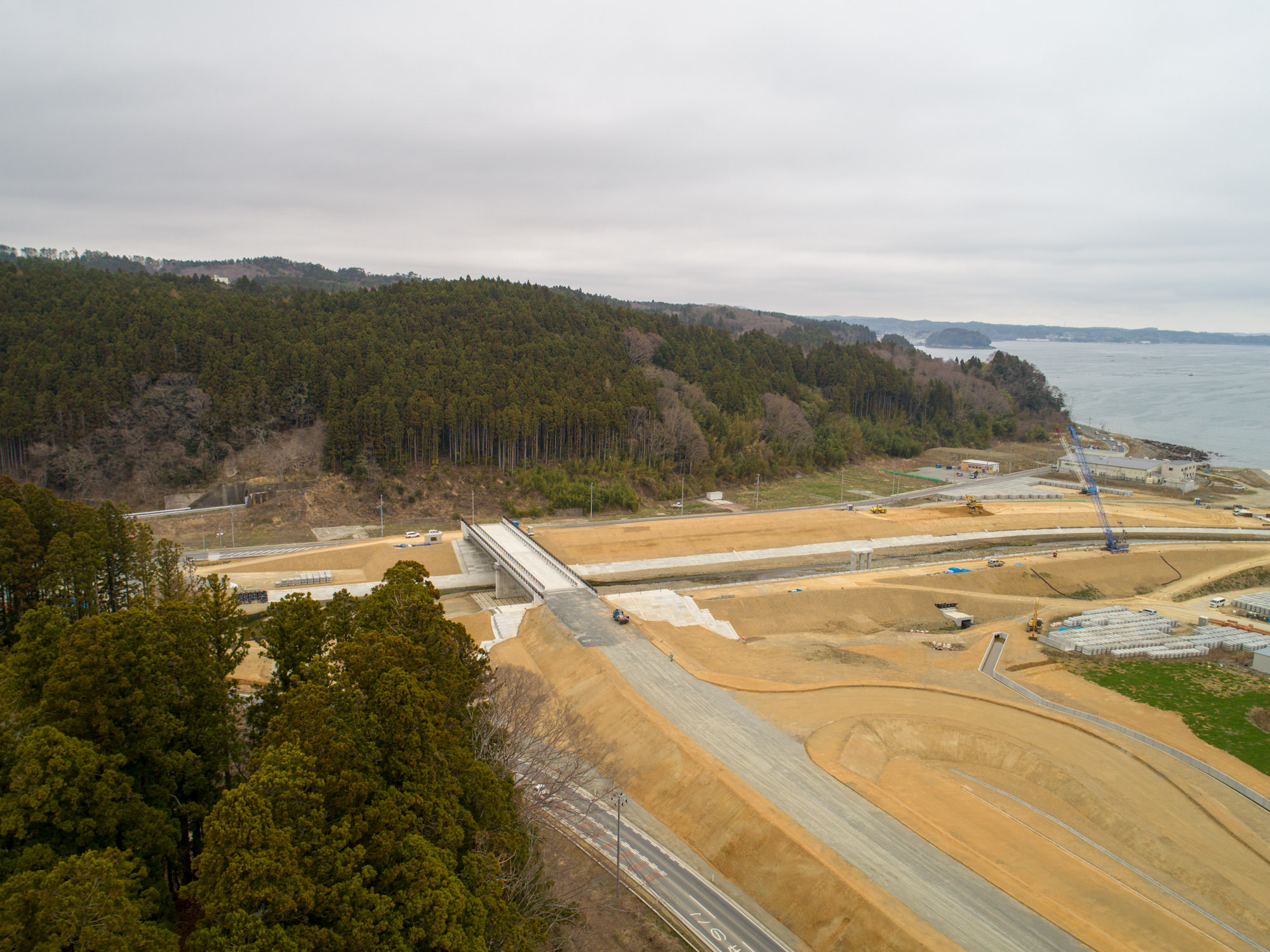 森林と海岸に近い場所で、複数の道路や通路が整備され、車両や建設機材が点在する工事現場を上空から捉えた写真で、整地された土地や建設途中の構造物、緑地が混在し、曇り空が広がる風景の写真