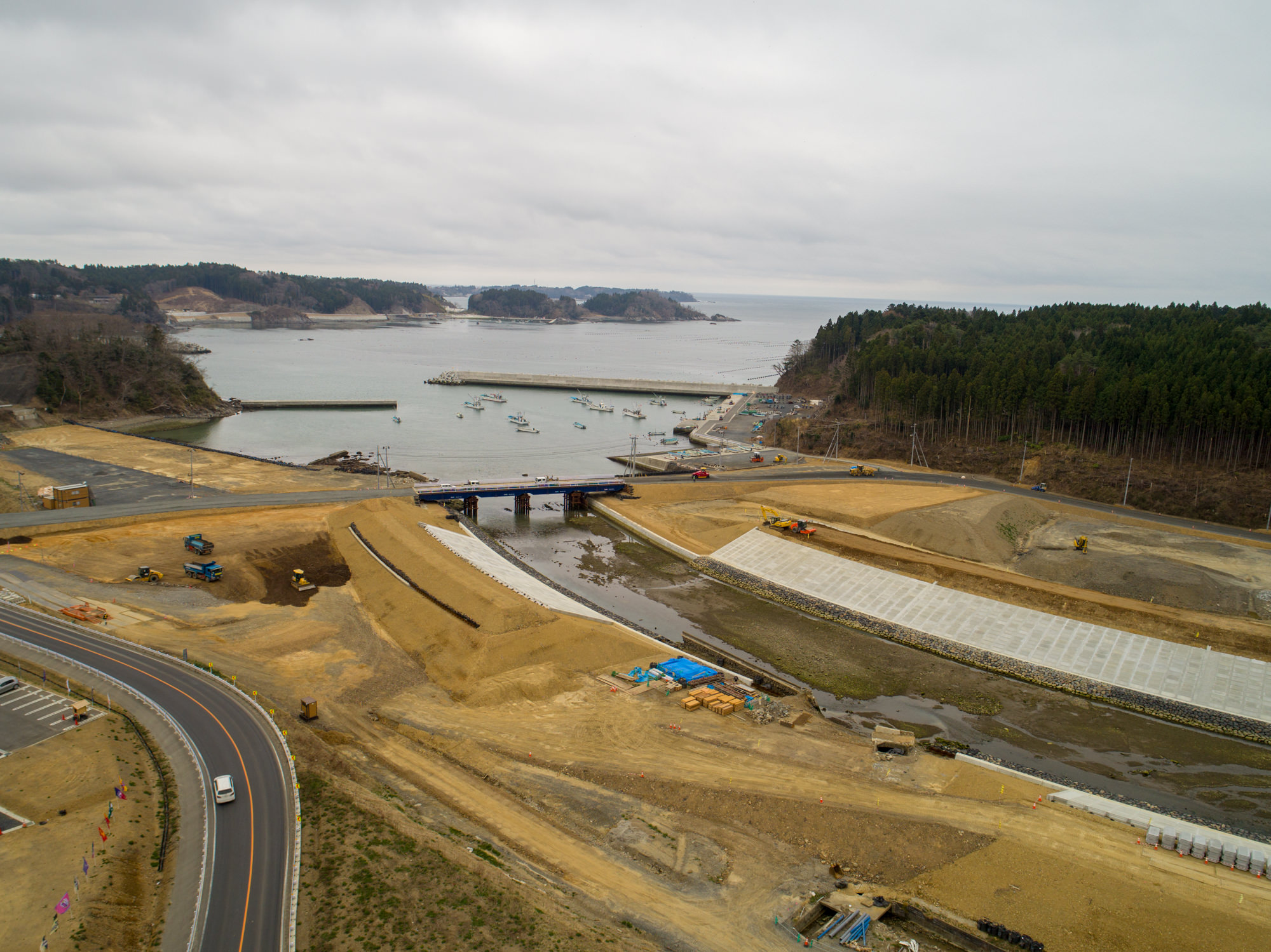 曇り空の下、港に複数の船が停泊する水辺を中心に、周囲で再開発または工事が進められている沿岸地域を上空から撮影したもので、橋が水路をまたいで港へと続き、道路や歩道が整備・改修されており、背景には森林や丘が広がる風景の写真