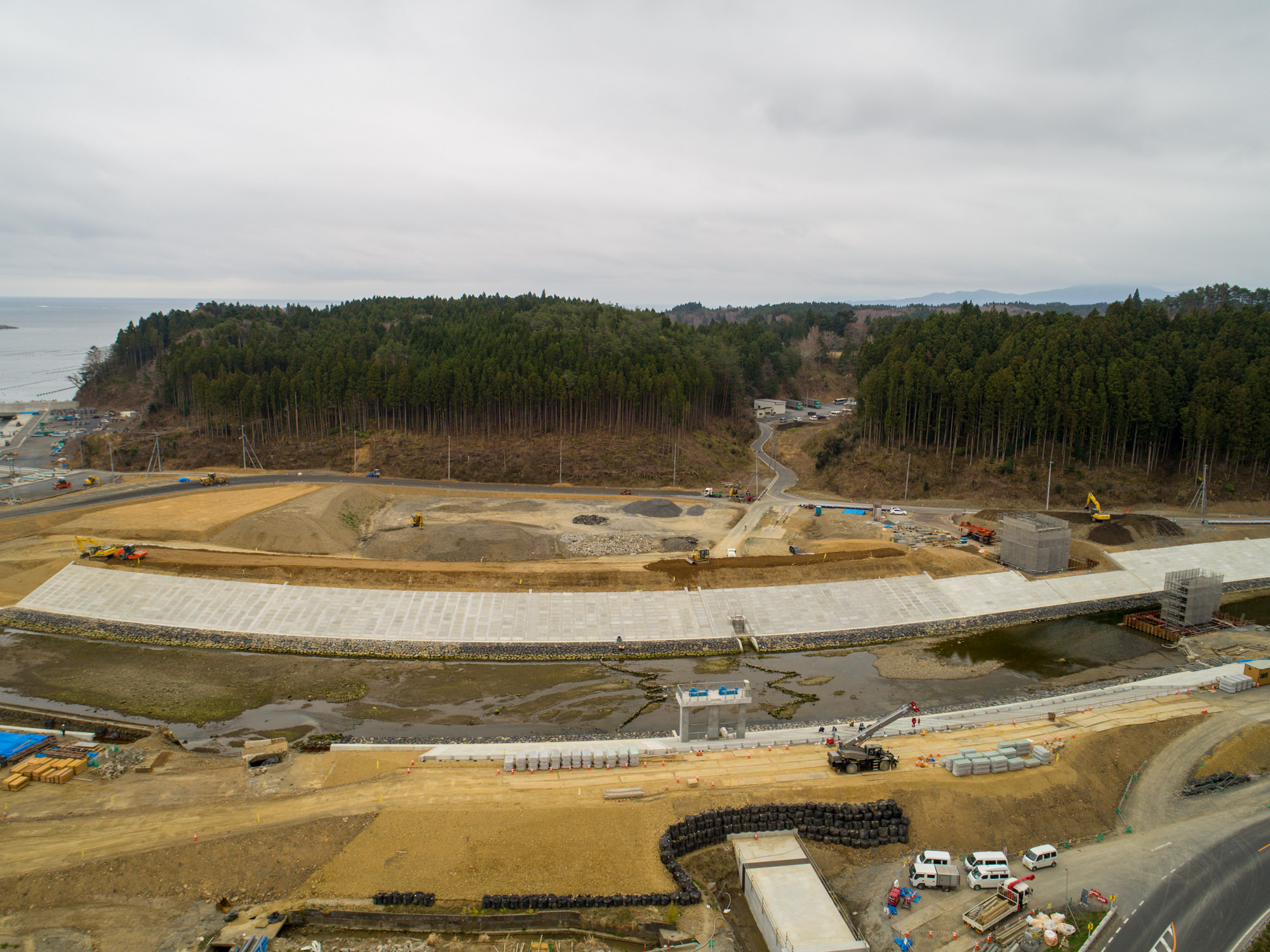 曇り空の下、前景にダムまたは防潮堤と思われる大きなコンクリート構造物があり、周囲には複数の建設車両や機材が点在し、背景には密集した森林へ続く道路が見える建設現場の風景の写真