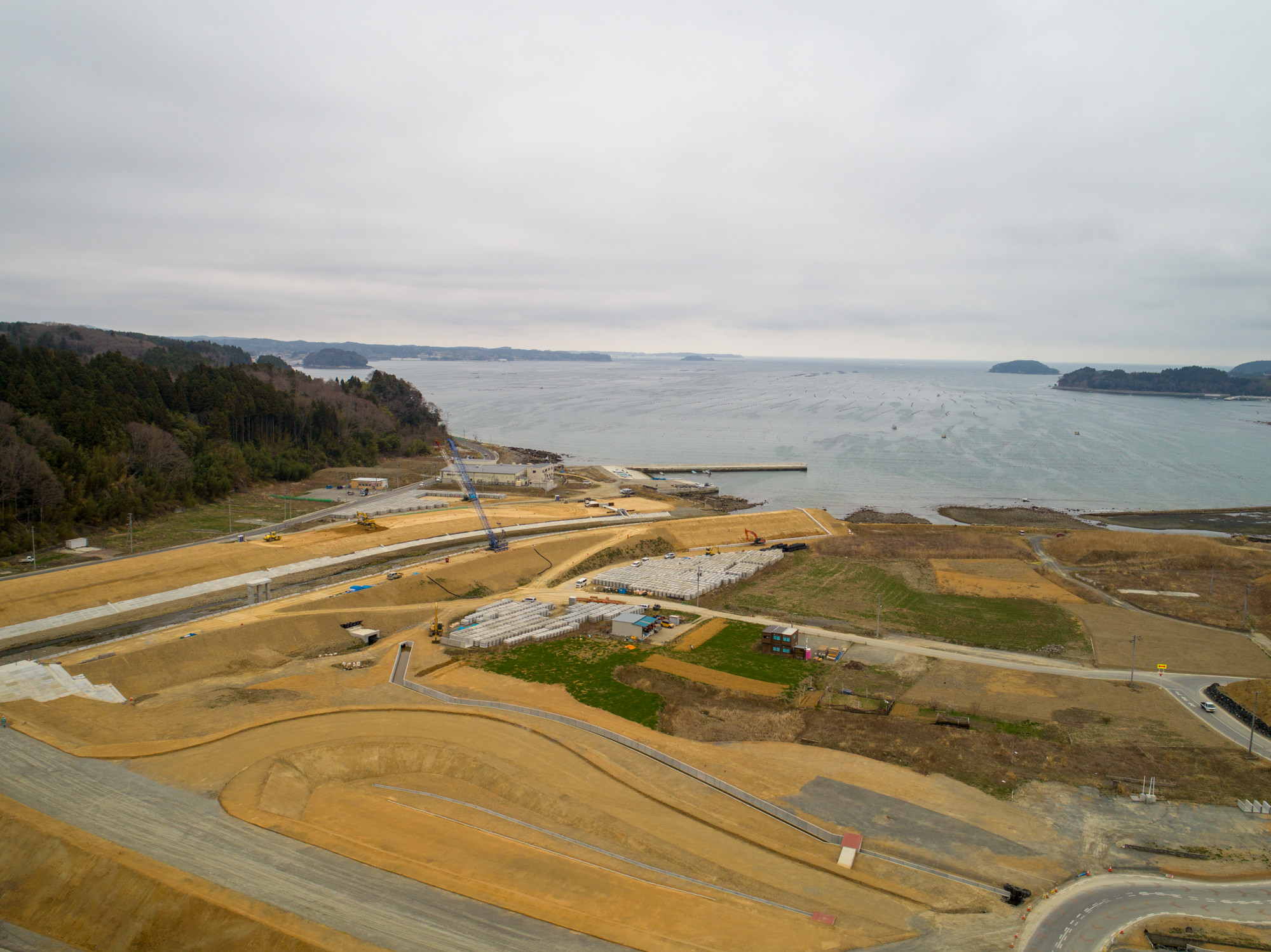 海に面した建設現場を上空から捉えた写真で、複数の道路や通路が新設または工事中であり、中央にはクレーンが見られ、工事が進行中であることが分かり、周囲には緑地や建物が点在し、背景には海と遠くの陸地が広がる風景の写真