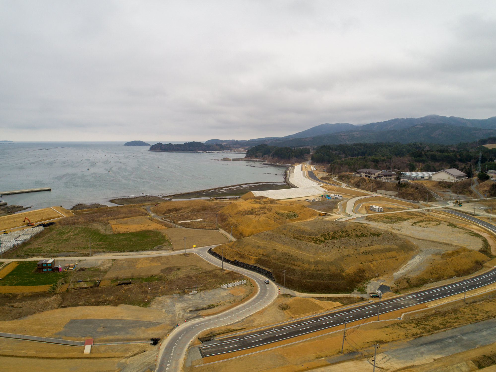 左側に海岸線が広がり、地平線まで続く海を背景に、舗装済みと工事中の曲がりくねった道路や通路が複数見られ、草地や裸地、建設機材が点在する地域を上空から捉え、奥には木々に覆われた丘や山が広がり、曇り空が空一面を覆っている風景の写真