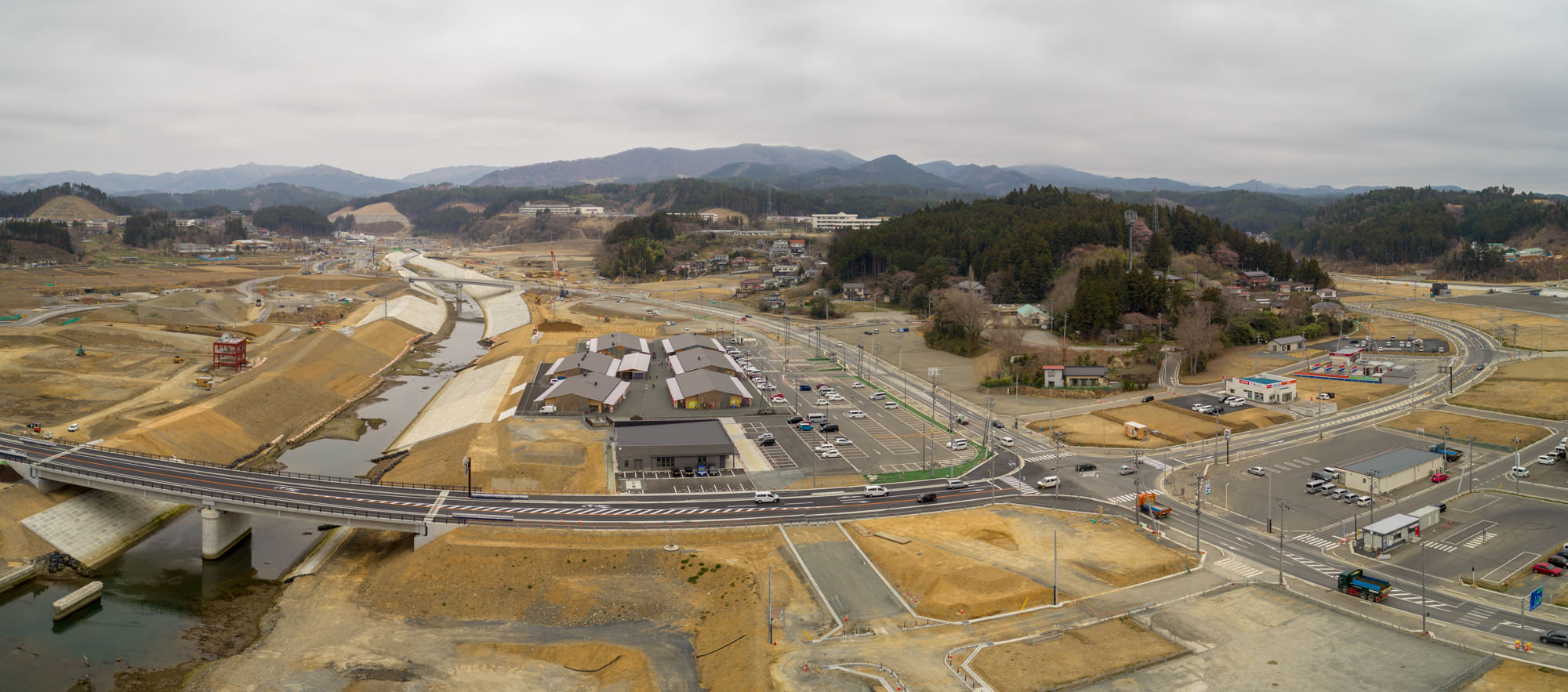 川に架かる橋と複数の道路が広がる地域を上空から撮影したもので、背景には山々が連なり、駐車場に車が並び、地域全体が開発または再整備中と思われる風景の写真