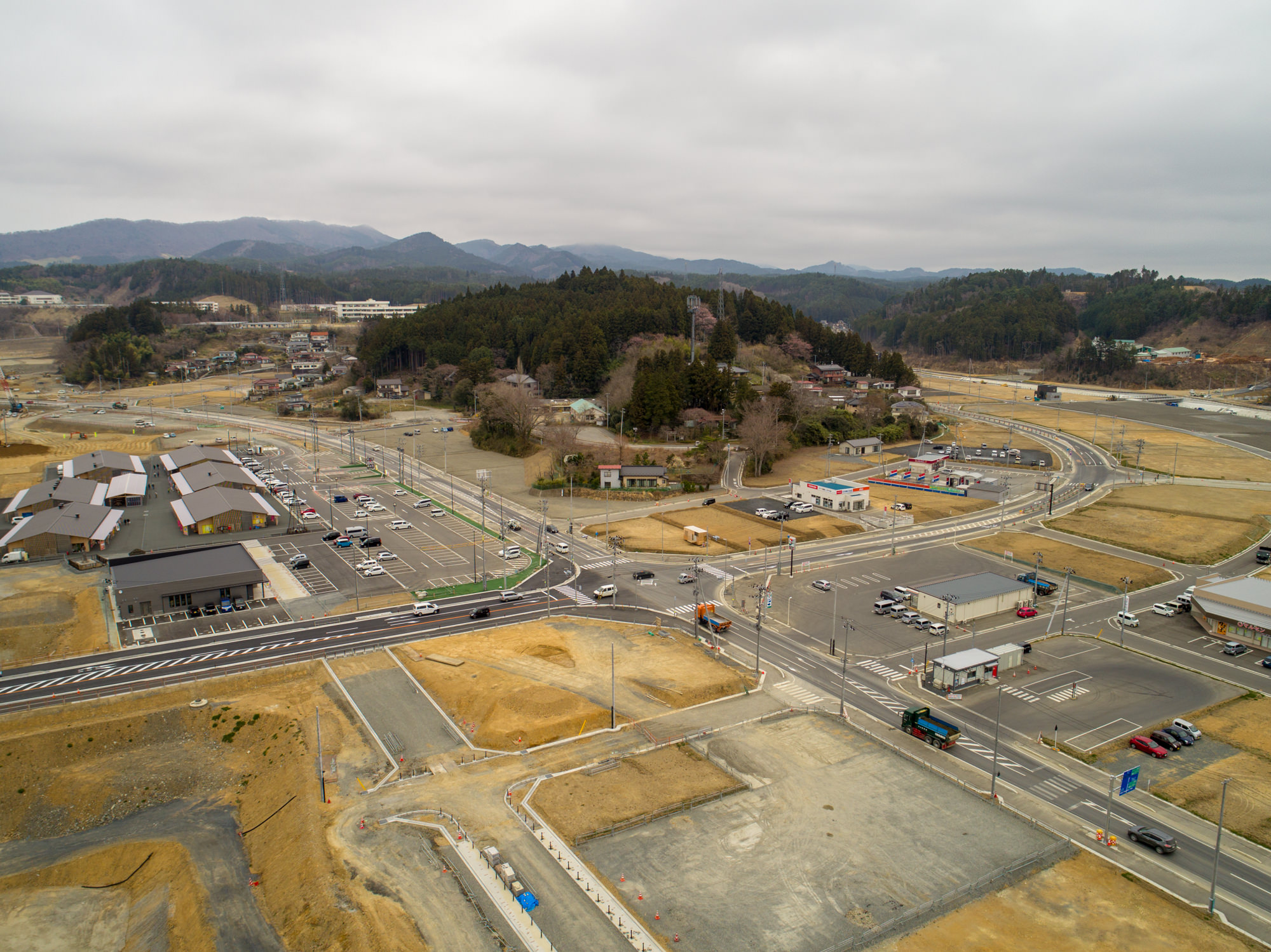 曇り空の下、小さな町または郊外地域を上空から撮影したもので、建物や道路、駐車場が点在し、車が複数見られ、背景には丘や森林、さらに遠くに山々が広がる風景の写真