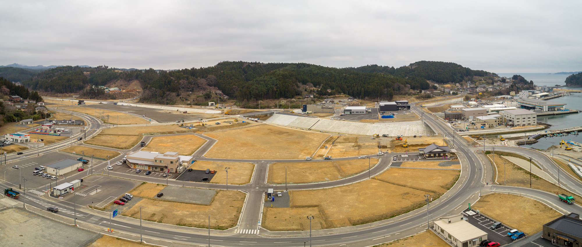 曇り空の下、右側に海が広がる沿岸地域を上空から撮影したもので、道路や建物、空き地が点在し、背景には木々に覆われた丘が見える、開発地と未開発地が混在する風景のパノラマ写真
