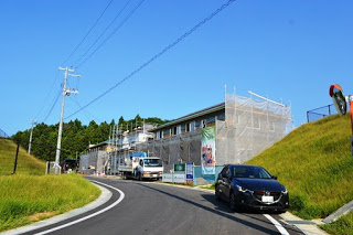 緩やかにカーブした道路の右側に1台の黒い車が停まった草の法面があり、その先に足場と保護ネットに覆われた建設中の建物があり、その前に1台の白い大型トラックが停まっている写真