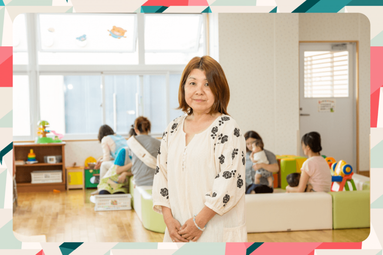 黒い花柄で白い服を着た女性が、明るい室内の子育て支援施設で子どもたちと保護者が遊ぶ背景の前に立っている写真