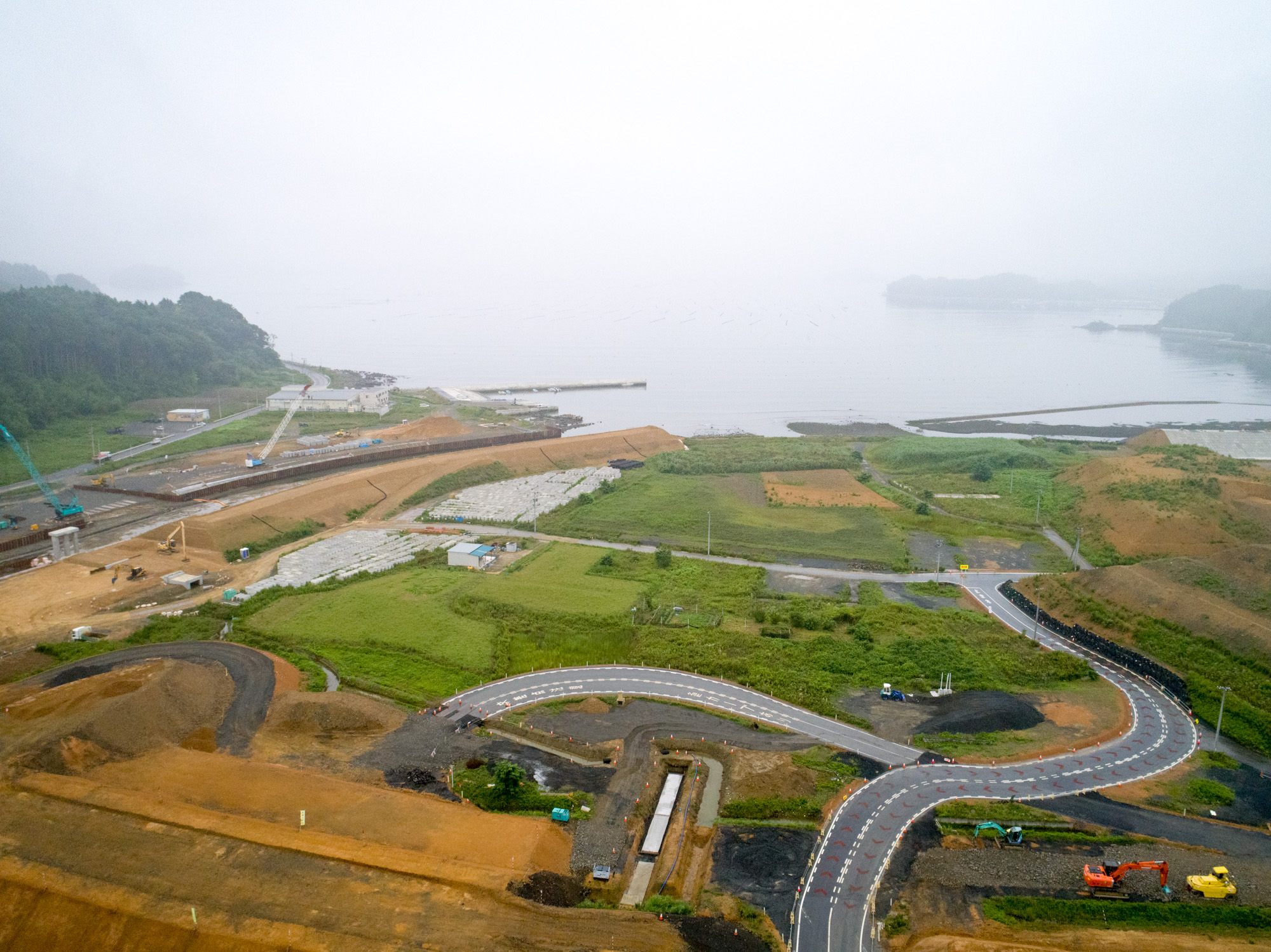 霧がかかった海沿いの地域で、曲がりくねった新しい舗装道路や建設機材が点在し、さらなる開発が予定されている土地が広がる、海と工事の様子が交差する風景の写真