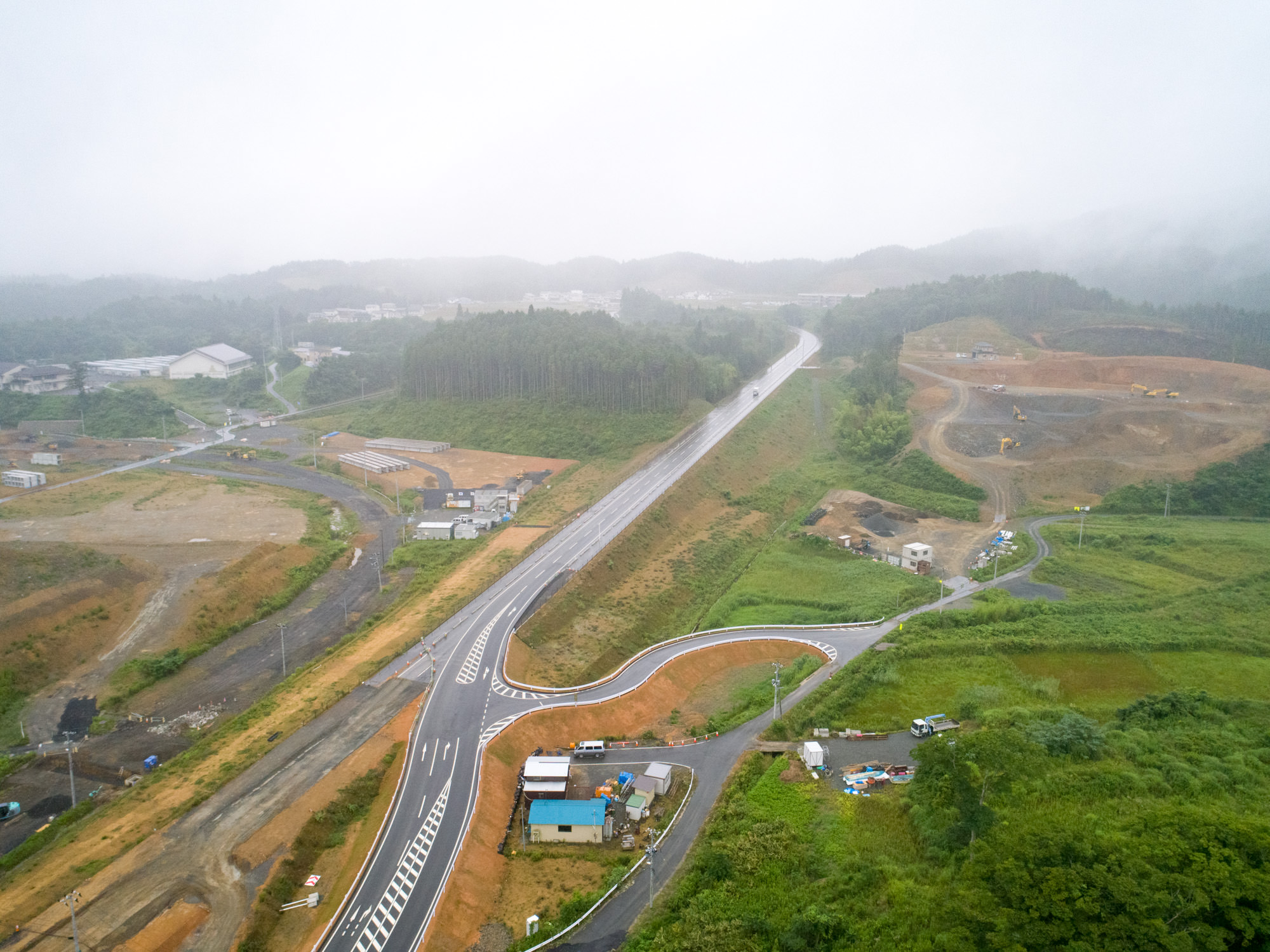 霧に包まれた丘を背景に、緑の田畑や整地された土地の中を多車線の新設道路が曲がりながら走り、交差点や建物も点在する、田園とインフラ整備が調和した風景の写真